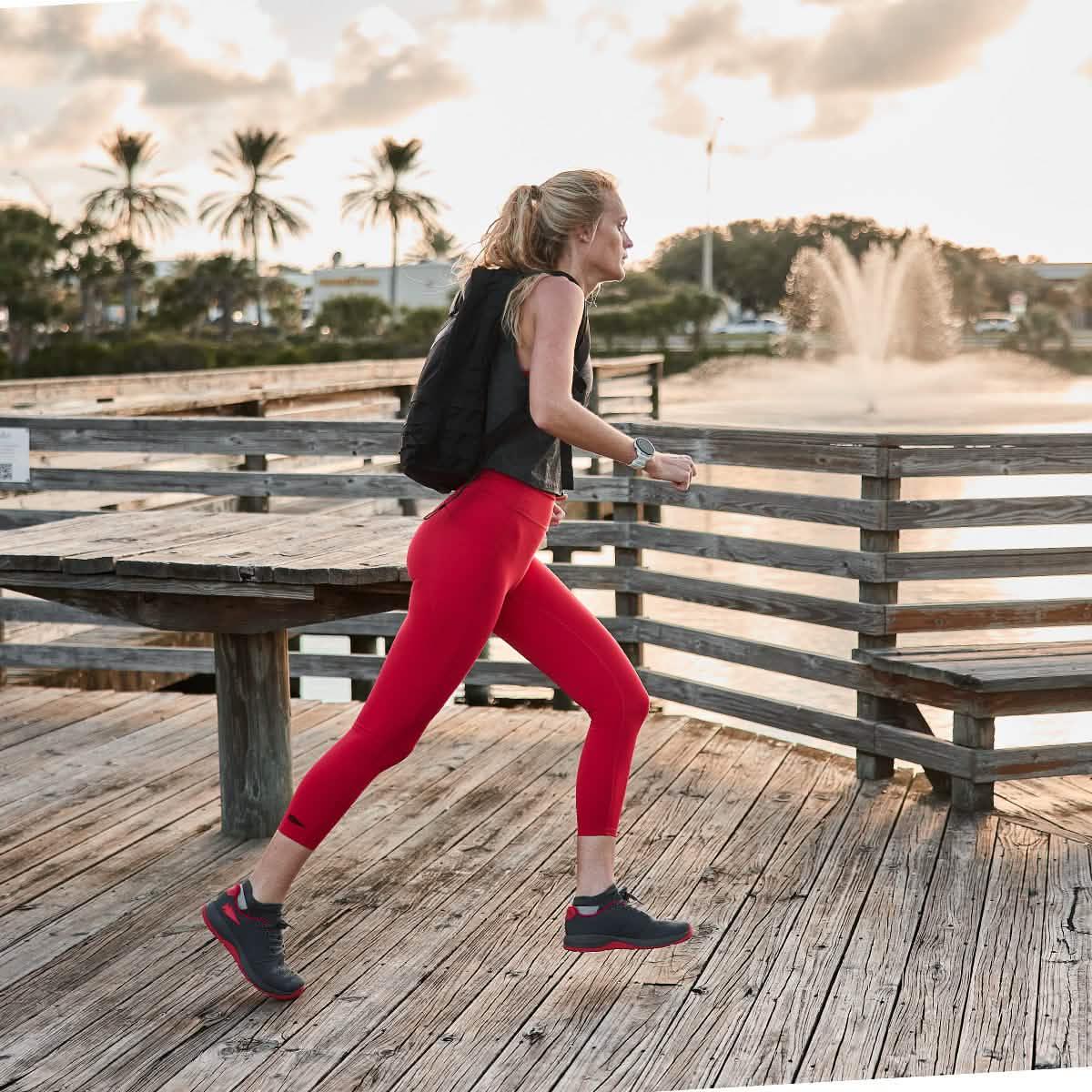 Woman running outdoors in GORUCK Wolf Grey and High Risk Red Ballistic Trainers, wooden deck, palm trees