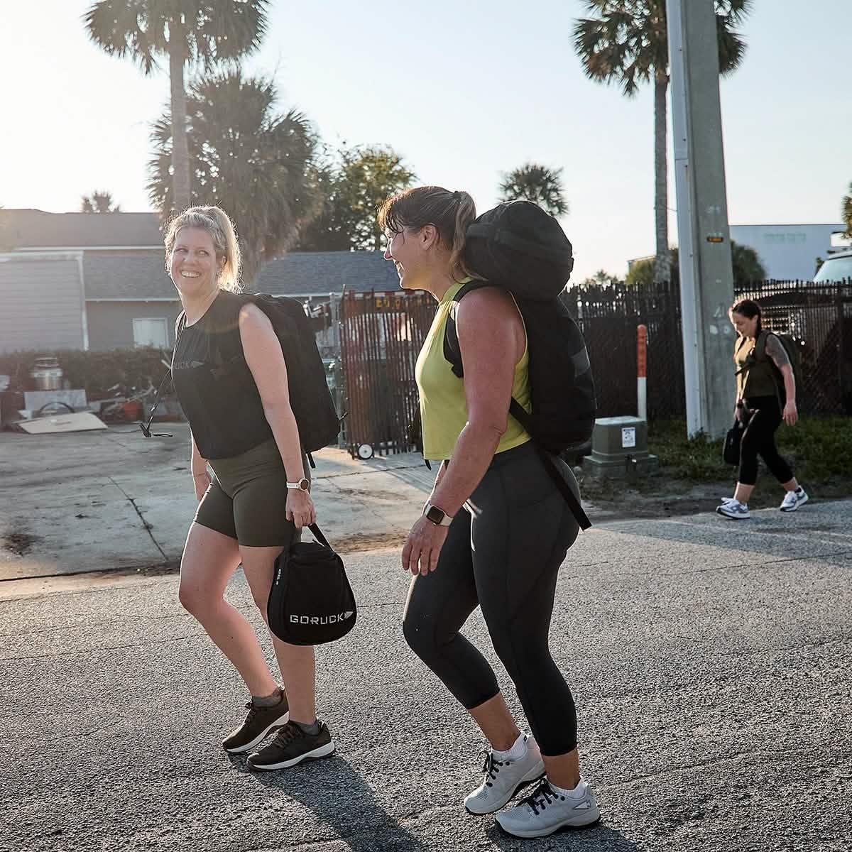 Two women walking outdoors wearing athletic gear and carrying GORUCK backpacks, smiling and enjoying the sun