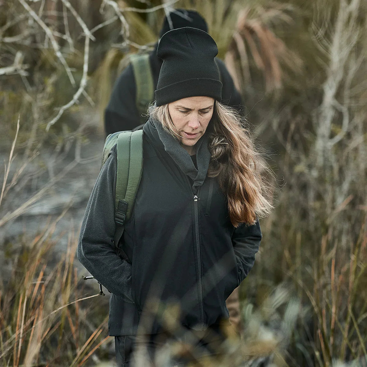 Woman wearing black fleece vest and beanie with green backpack hiking through dense brush outdoors