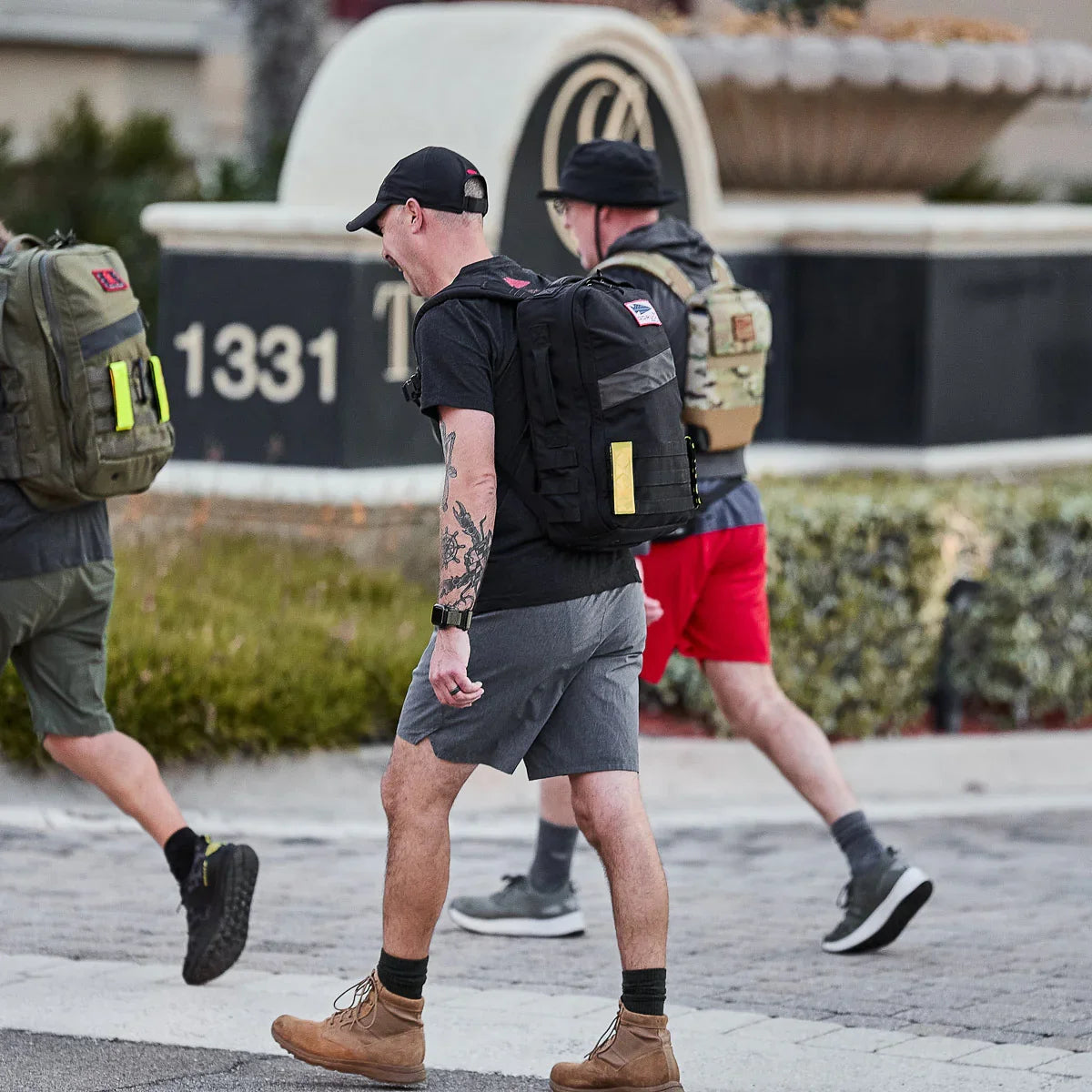 Three men walking outdoors wearing tactical rucking backpacks and casual gear on a paved path