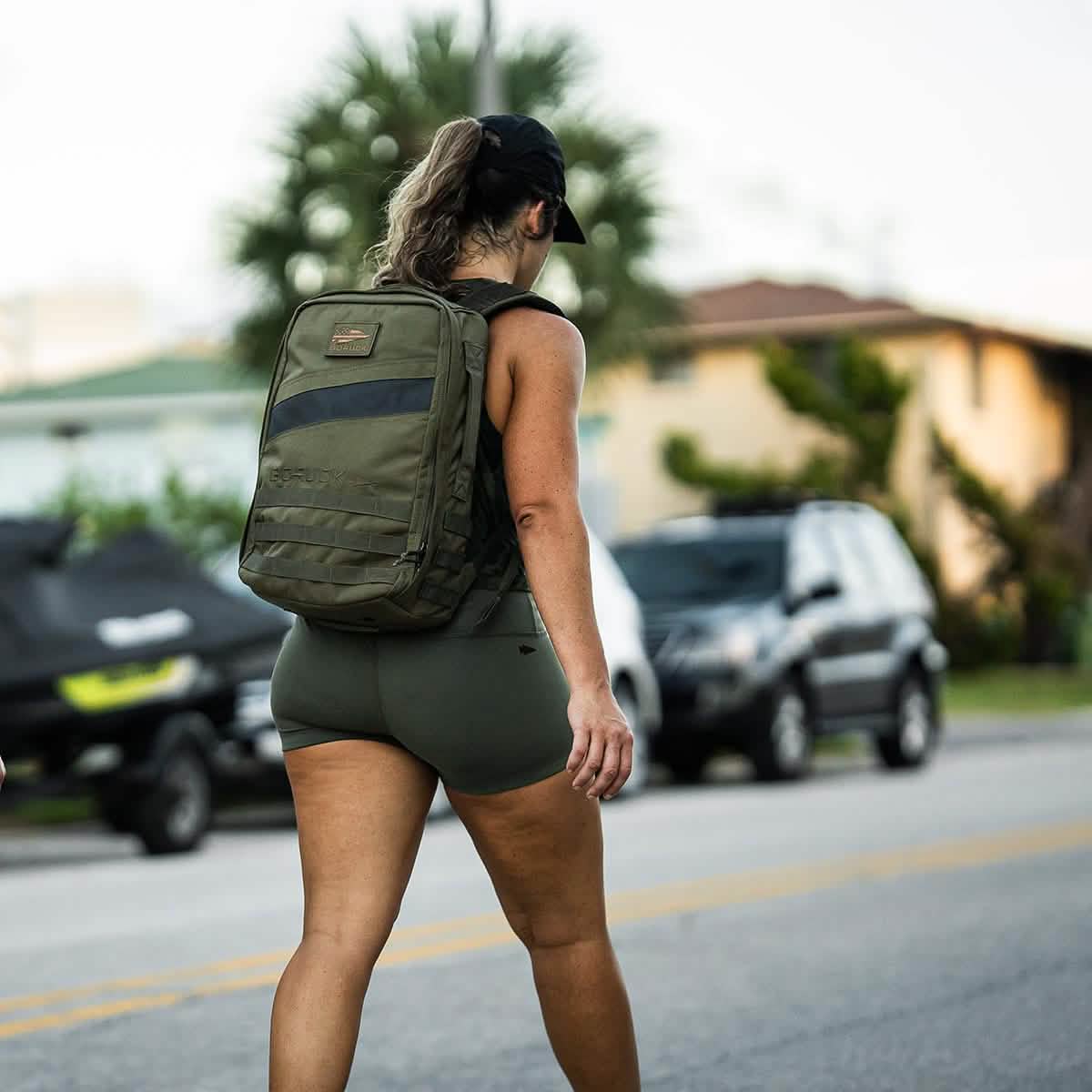 A woman in GORUCK’s high-waisted Women’s Squat Shorts made with durable ToughFlex Fabric and a cap walks down a street with a green backpack. Palm trees and vehicles are visible in the background, emphasizing her relaxed stride.