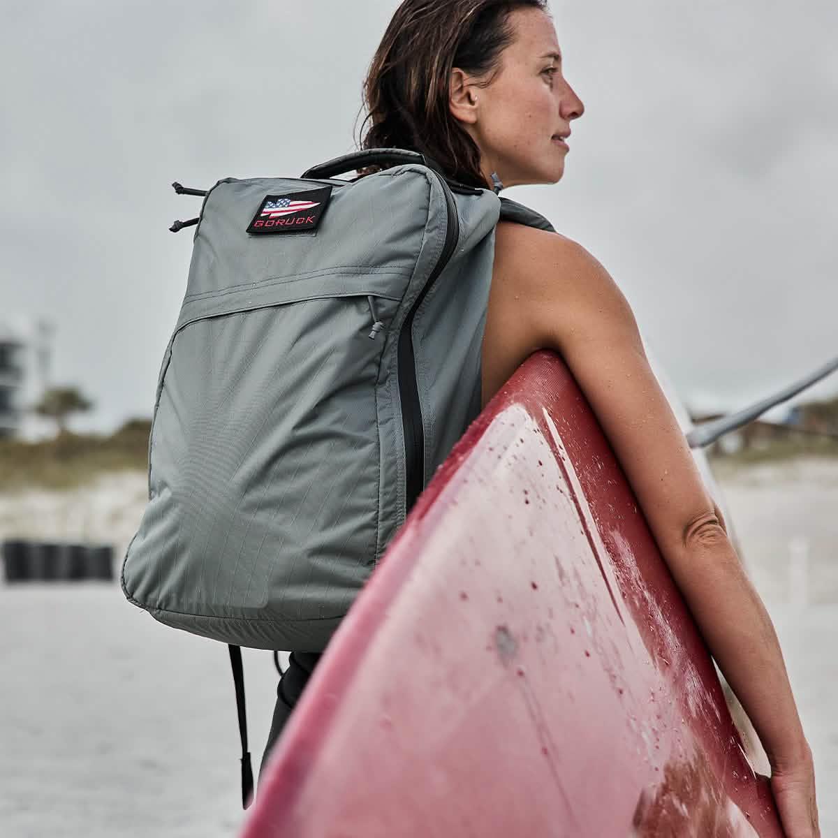 Woman carrying a gray GORUCK backpack with American flag patch, holding a red surfboard on a beach