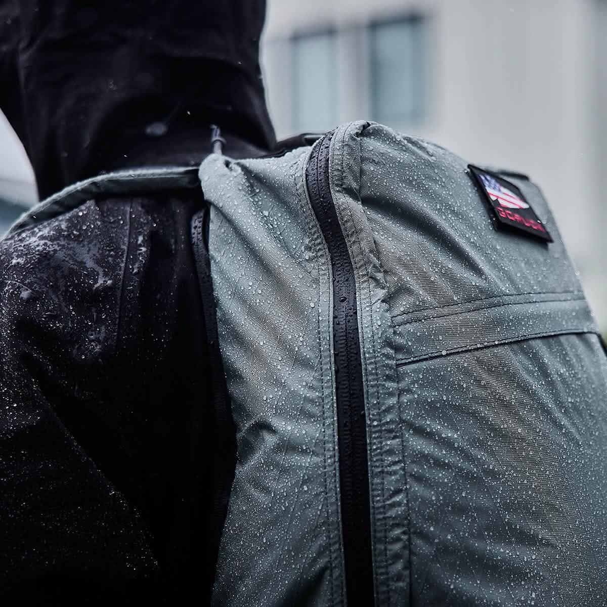Close-up of waterproof GORUCK rucksack with water droplets on fabric, worn over black jacket in rainy setting
