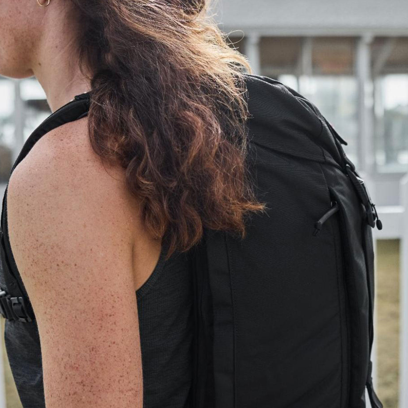 A person with curly brown hair wears a black backpack outdoors, shown from the side.