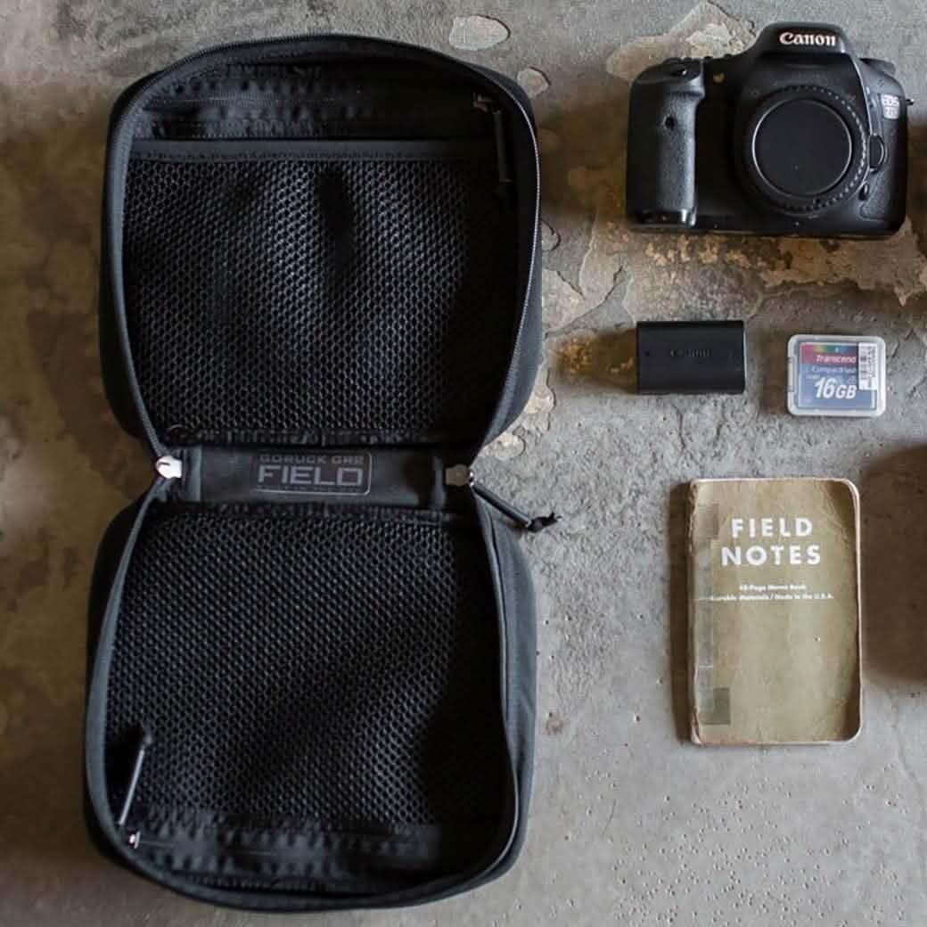 Open black GORUCK GR1 Field pouch next to Canon EOS 7D camera, battery, 16GB SD card, and Field Notes notebook on concrete