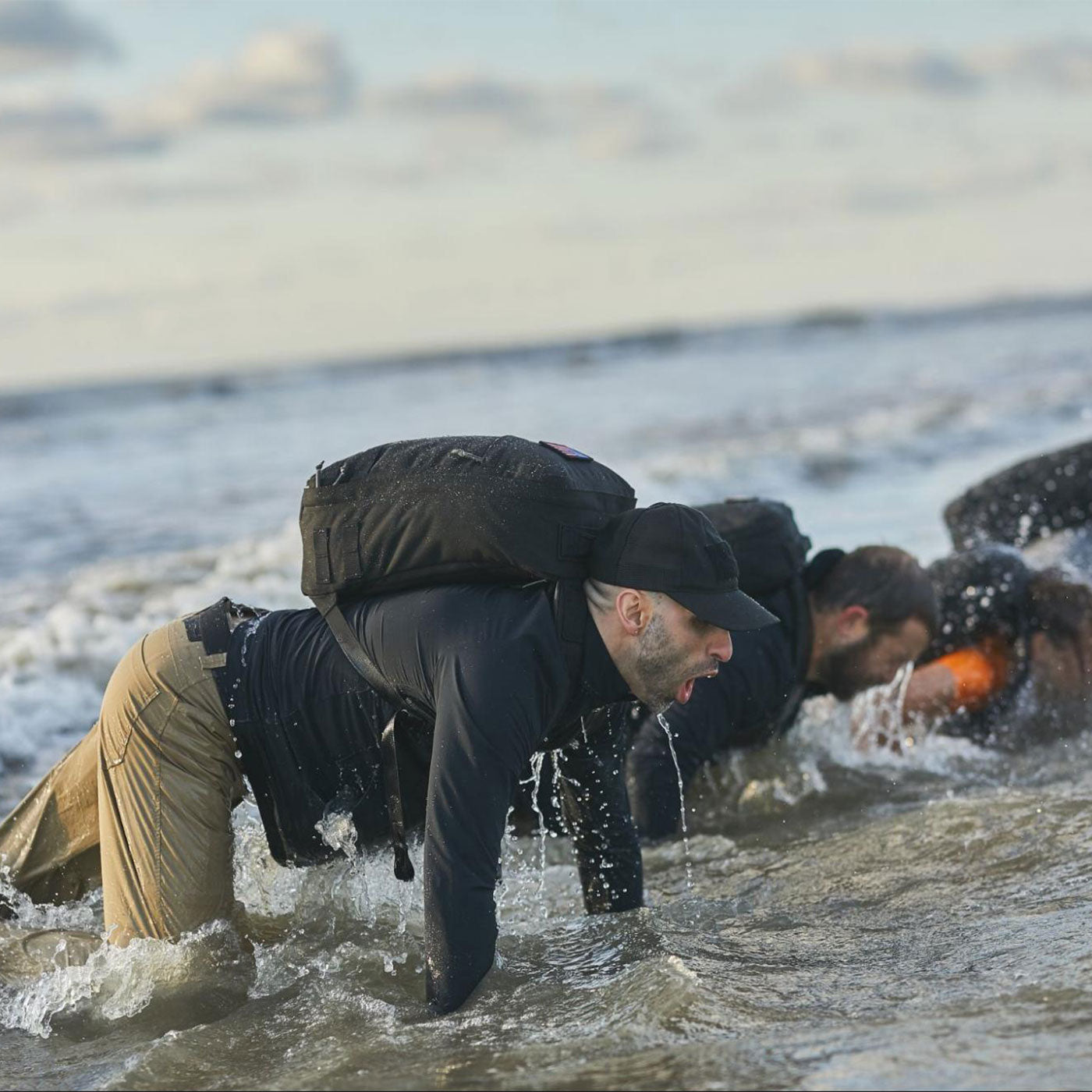 Men wearing backpacks crawl through shallow ocean water, splashing as they move forward.