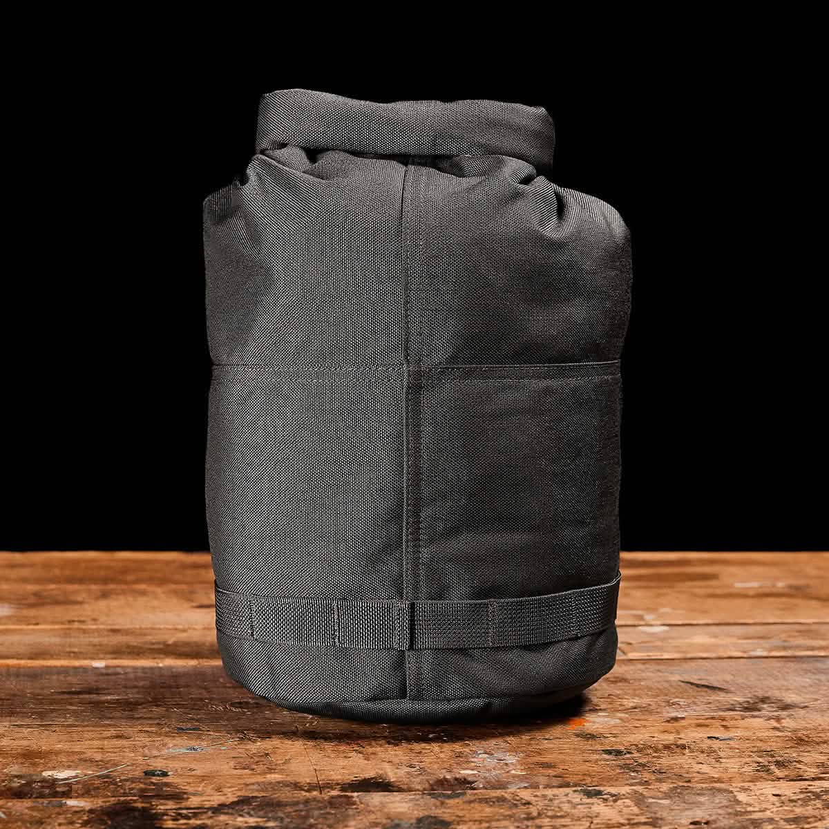 A high-key image showcases the GORUCK Brick Bag (5-50 LBS), a dark green, cylindrical canvas accessory with a rolled top that evokes military gear. It is positioned on a rustic wooden surface against a black background, featuring visible stitching and a strap near the base.