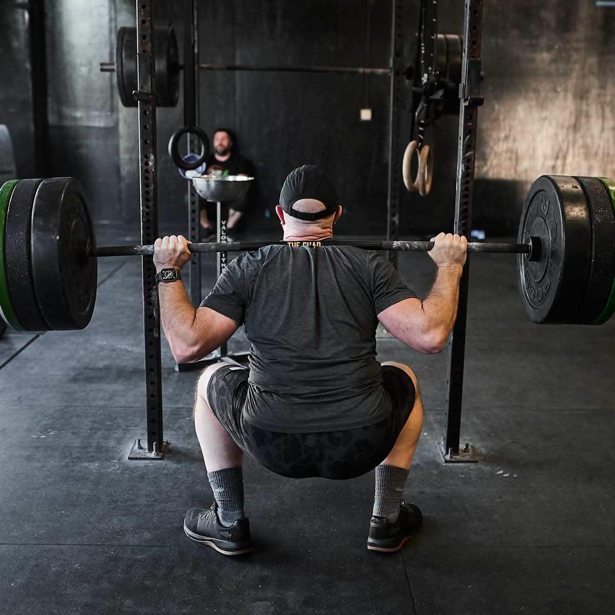 Man wearing black cap and athletic clothes performing heavy barbell squat in gym with black walls and workout equipment
