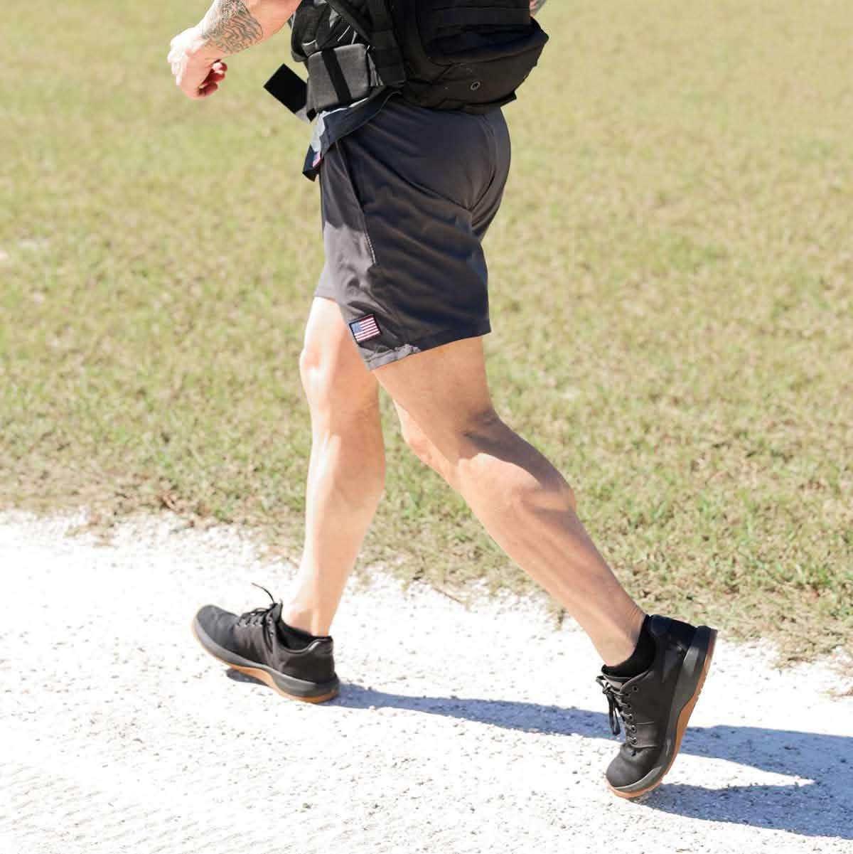 Person running outdoors wearing black athletic shorts with American flag patch, black shoes on grass and gravel