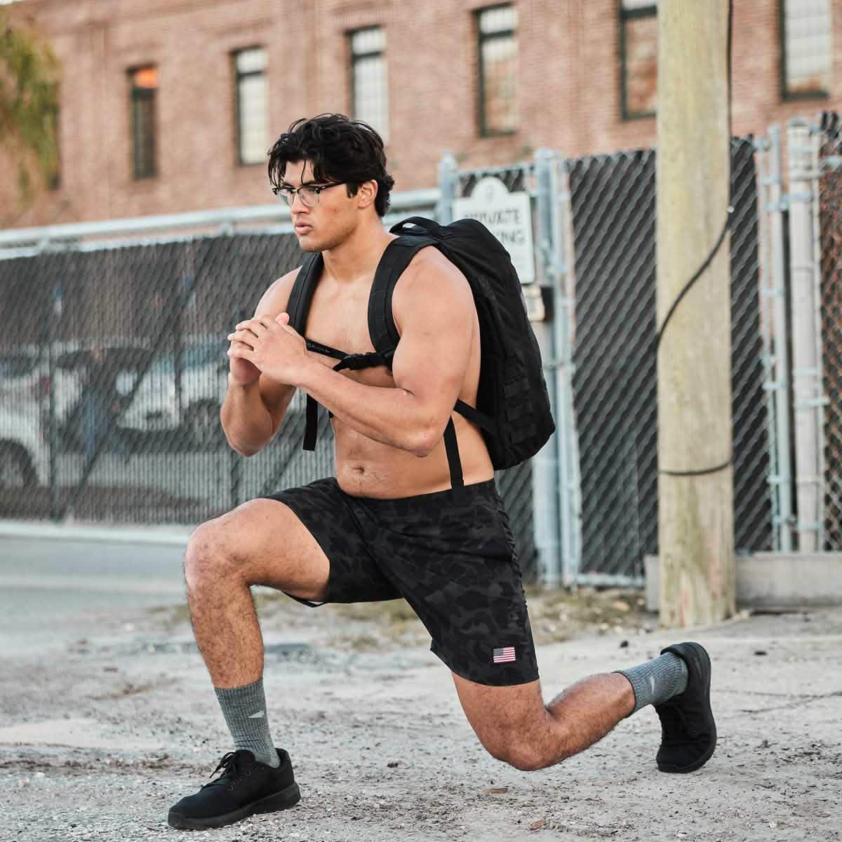 Shirtless man wearing glasses and black shorts with US flag patch, doing lunge exercise with black GORUCK backpack on urban street