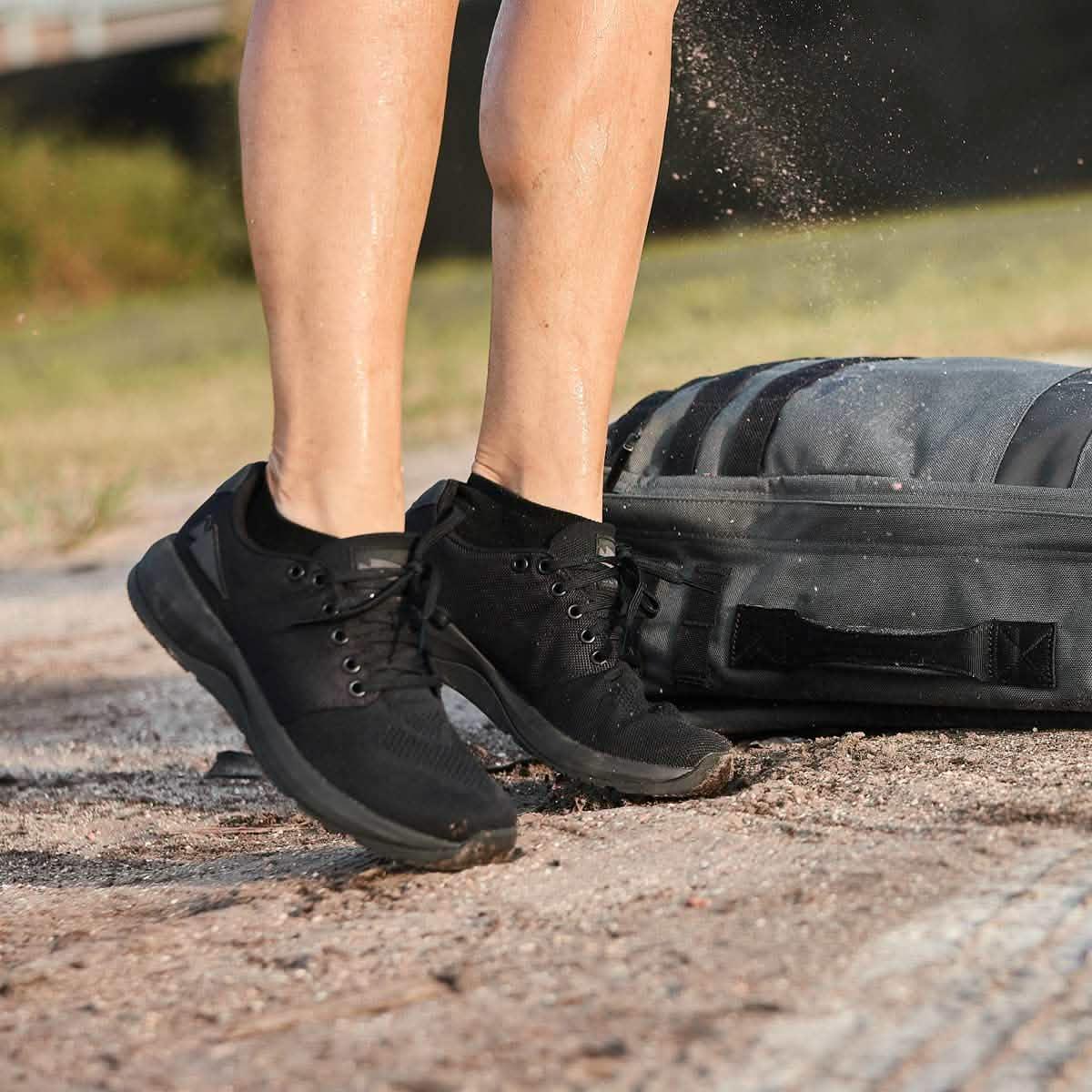 Person wearing black athletic shoes jumping outdoors near a black tactical rucksack with dust flying