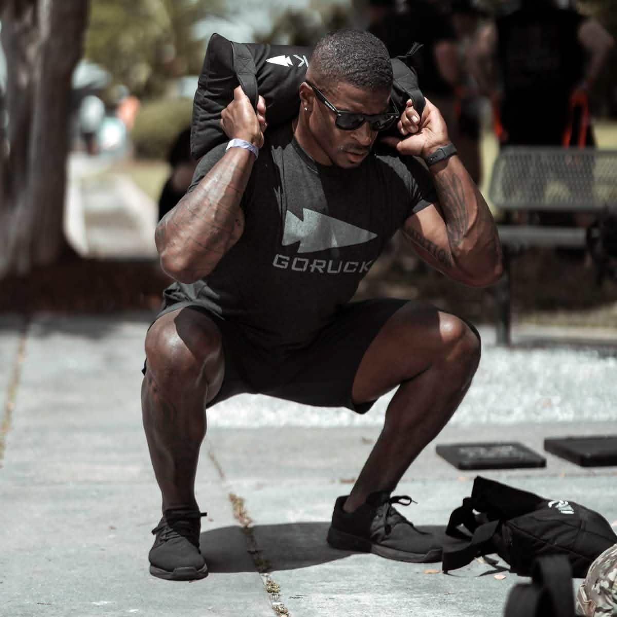 Muscular man wearing GORUCK shirt and sunglasses performing weighted squats outdoors with a rucksack