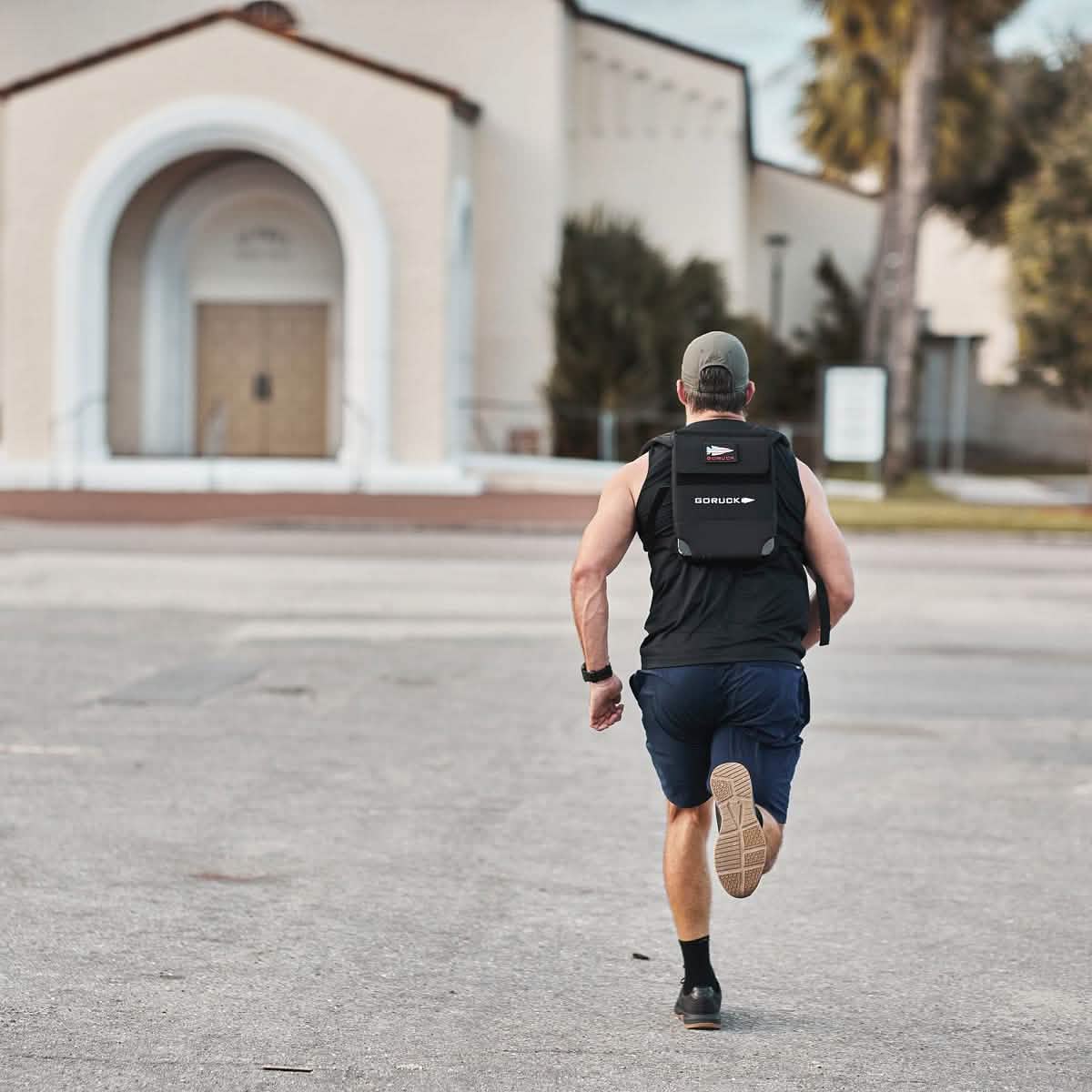 A person jogs along a paved path in GORUCK's Men's Ballistic Trainers in Black + Gum with a Black Reflective Spearhead, moving towards a building with an arched entrance. They're equipped with a backpack, shorts, a sleeveless top, and a cap. In the background, trees and grass are visible.
