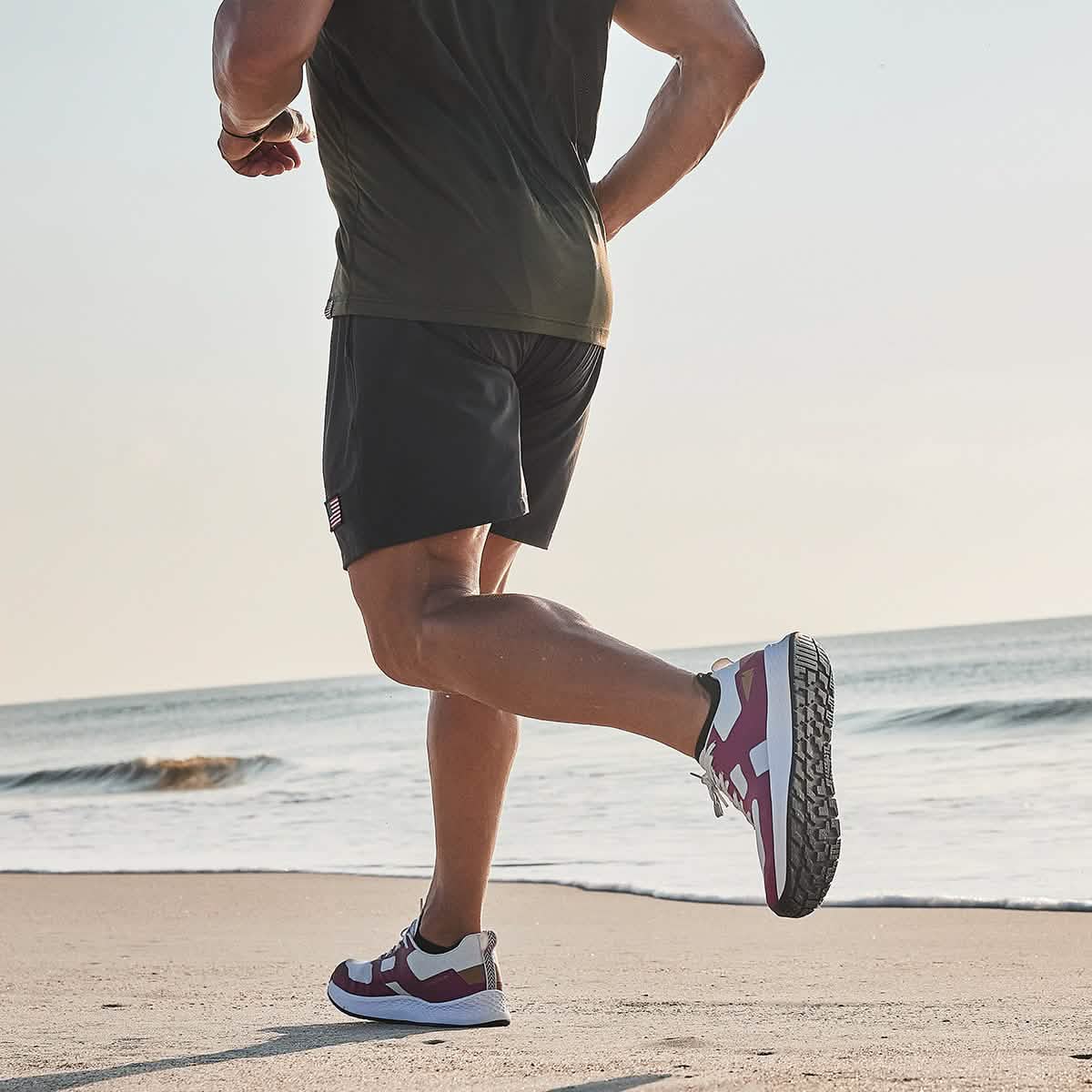A person, embodying the spirit of a Men's Rough Runner - Baton Rouge by GORUCK, sprints along the beach with waves crashing in the background. They wear a dark green shirt, black shorts, and maroon and white sneakers. The clear sky hints at a sunny day perfect for such Special Forces-like endurance.