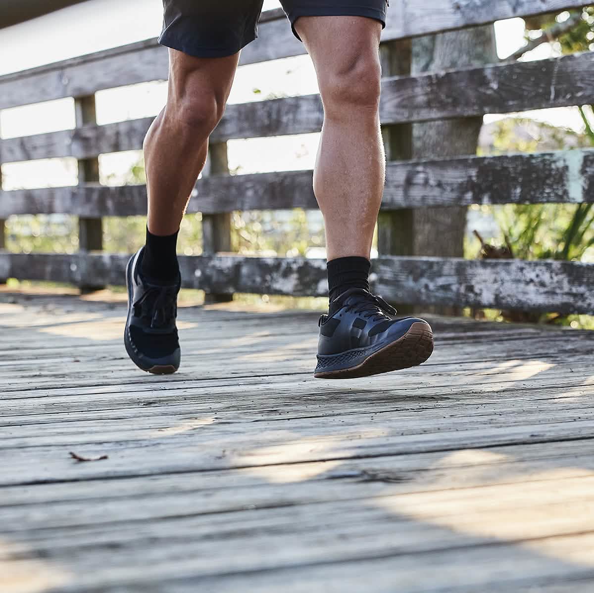 A person wearing the Men's Rough Runner - Black + Gum athletic shoes by GORUCK, featuring a gradient density EVA midsole, and shorts walks on a wooden boardwalk. The morning sun casts shadows on the planks, and a weathered fence lines the path.