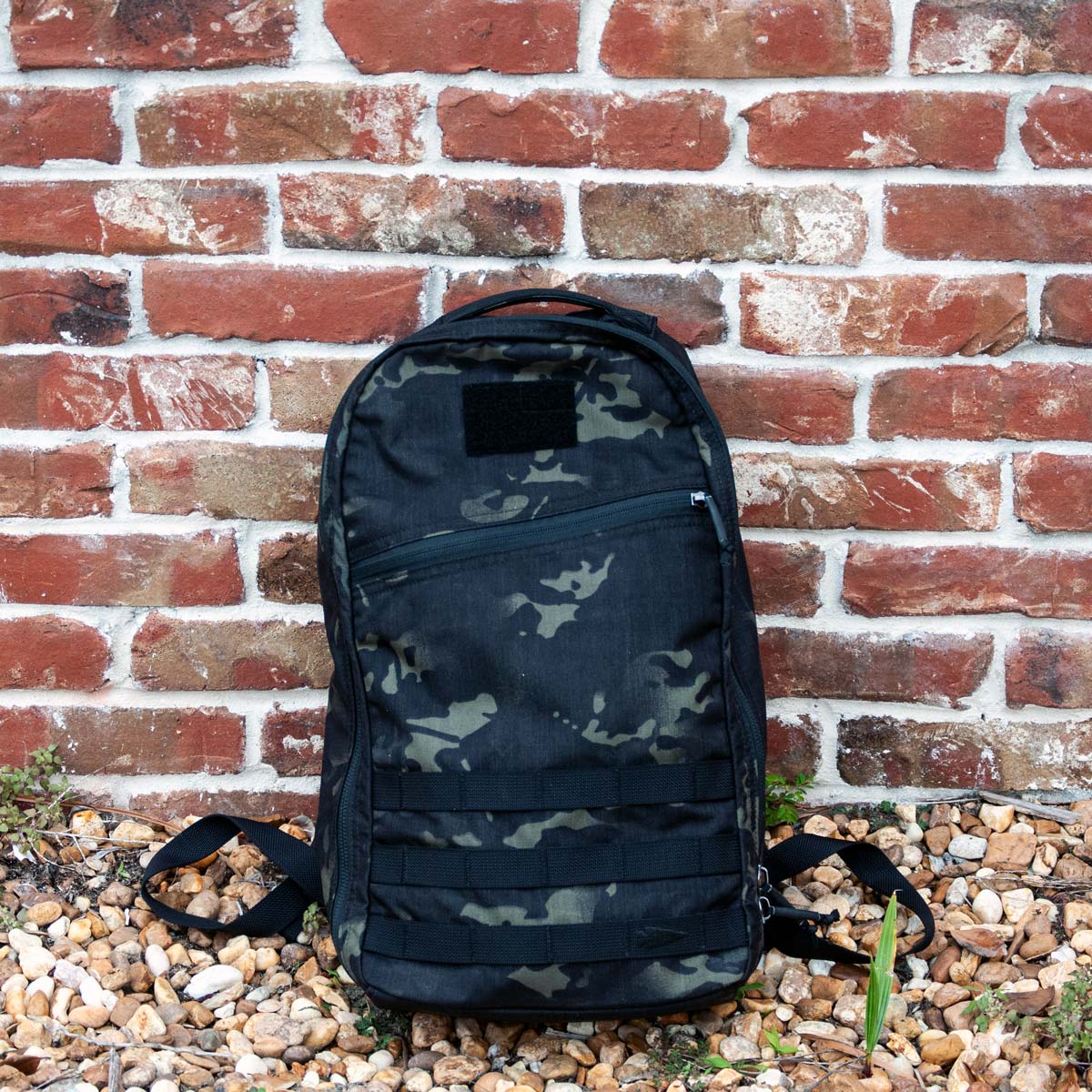 The Bullet Ruck Classic - Ballistic Nylon Cordura backpack in black camouflage rests on gravel before a red brick wall.