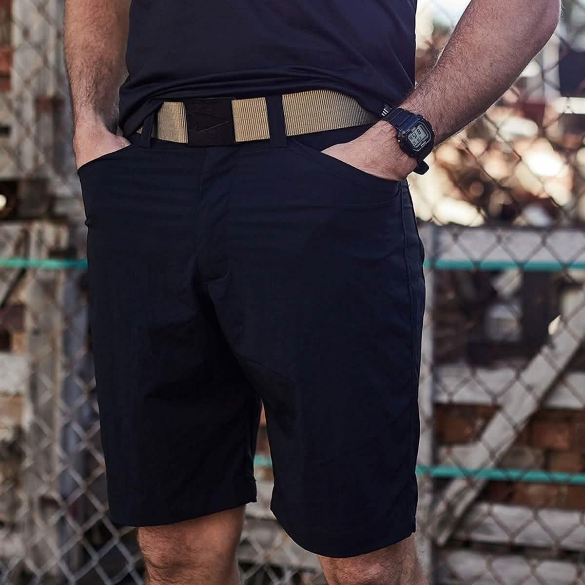 Man wearing black tactical shorts with a tan belt and digital watch standing near chain link fence