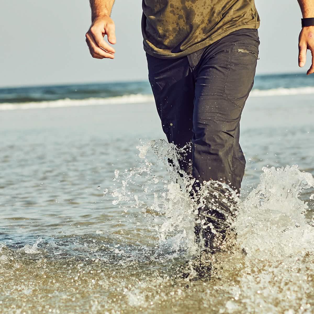 Person walking through shallow ocean water wearing black GORUCK tactical pants and a wet olive shirt