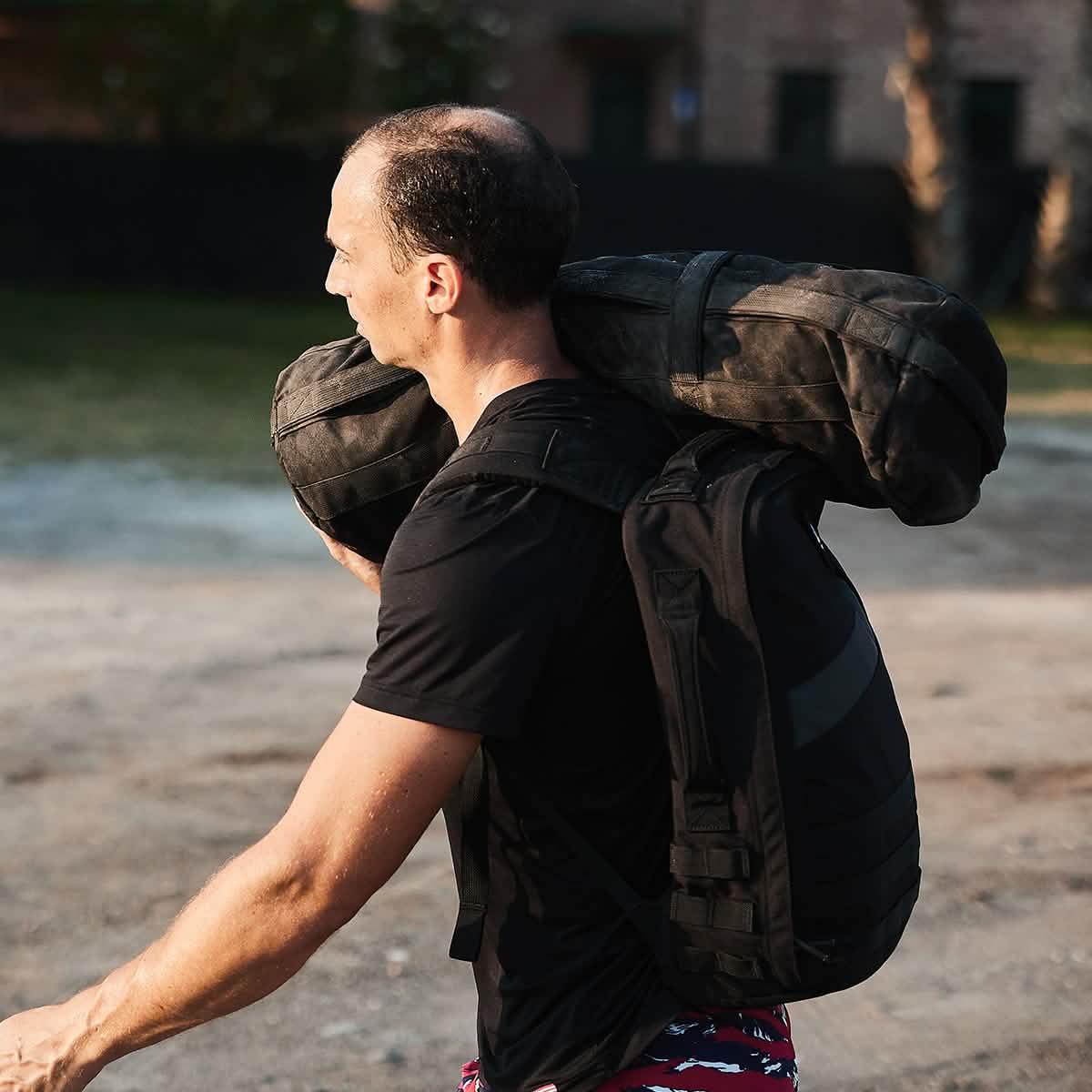 Man wearing black shirt carrying large black GORUCK sandbag on shoulder in outdoor setting