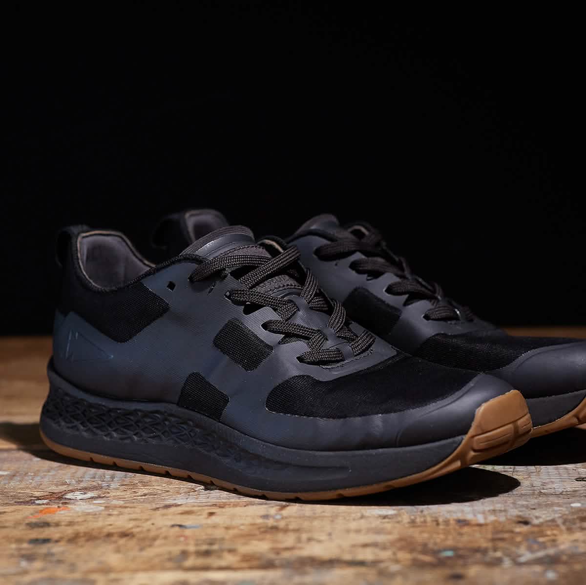 A pair of Men's Rough Runner - Black + Gum shoes from GORUCK rests on a wooden surface against a dark background. Featuring black athletic styling and brown soles, these shoes boast a textured Gradient Density EVA Midsole and sleek black laces, making them ideal for any challenging terrain.