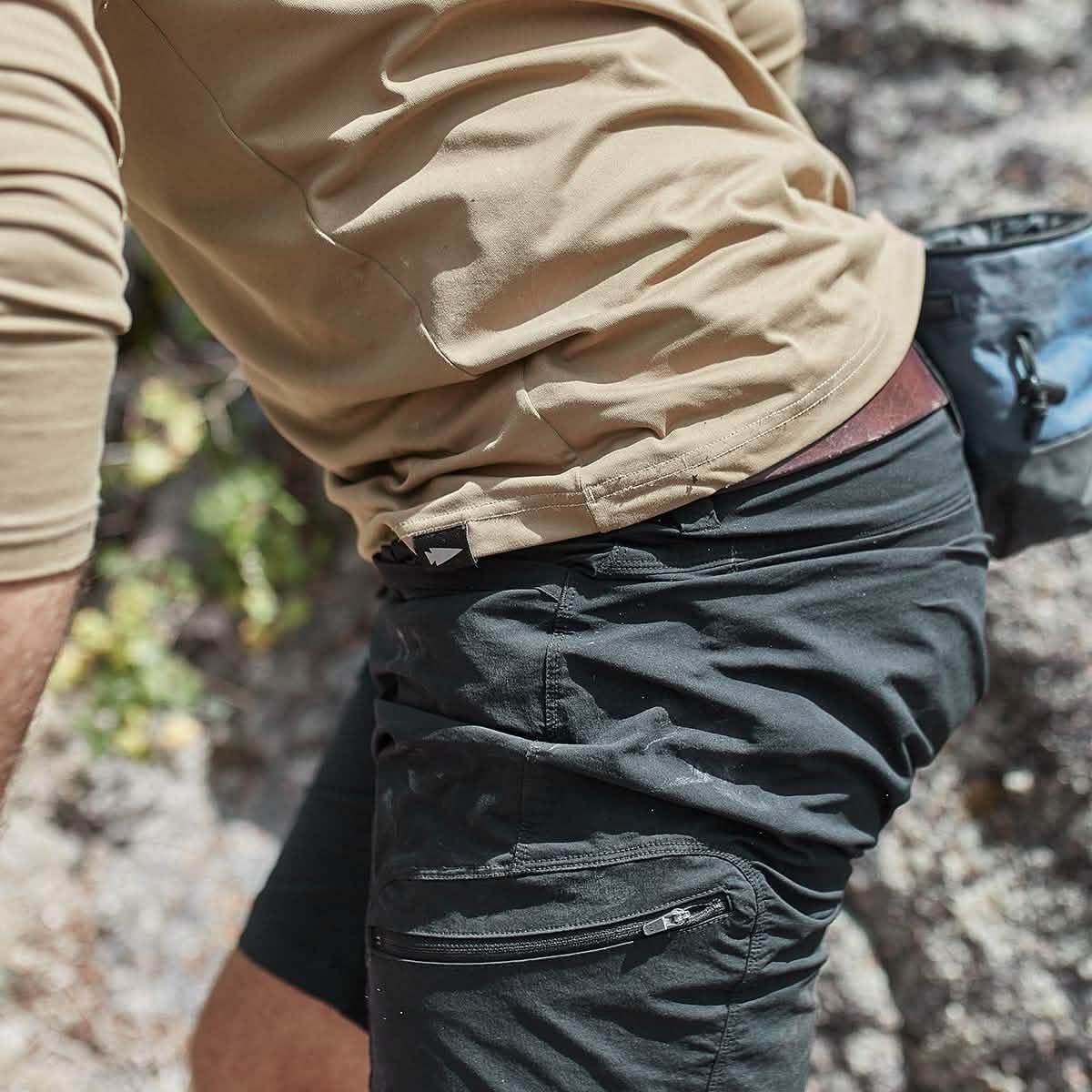 Person wearing black tactical shorts with zipper pocket and tan shirt outdoors on rocky terrain