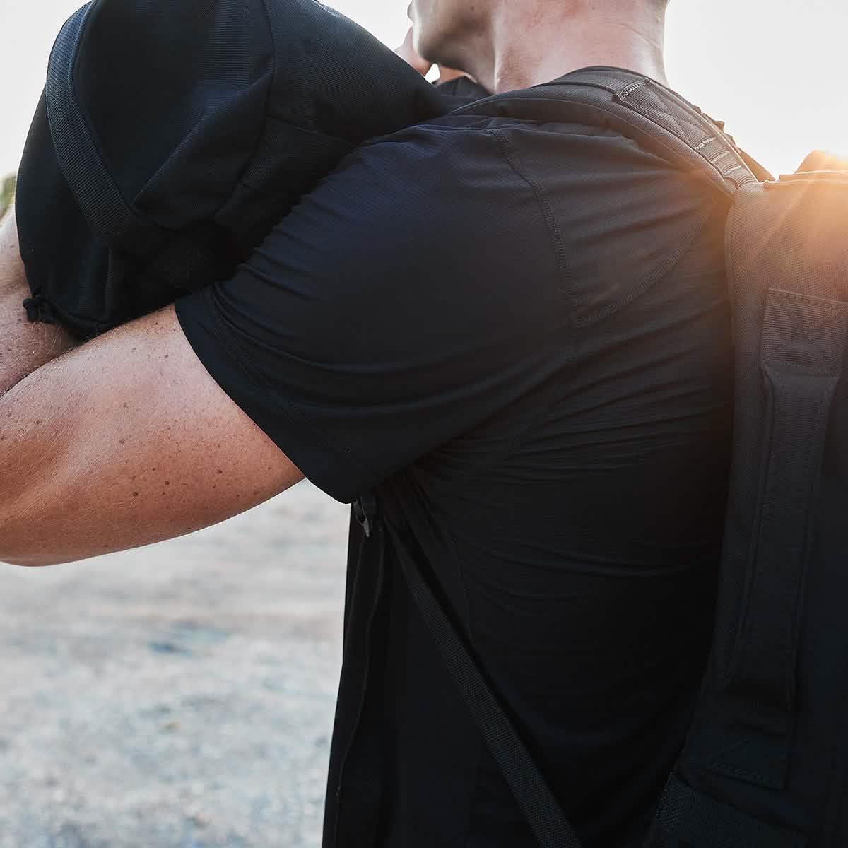 Man wearing black tactical rucking gear carrying a weighted sandbag outdoors with sunlight in background