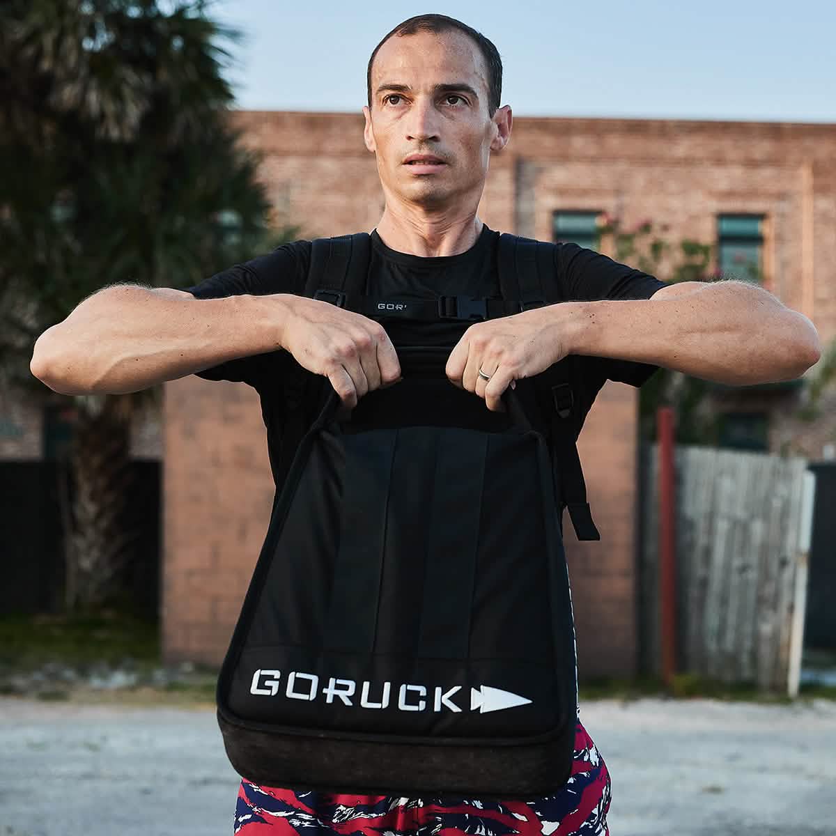 A man outdoors wears a black GORUCK backpack and the Men’s USA Performance Tee - ToughMesh, a featherweight, quick-drying training shirt, with a brick building and palm tree in the background.