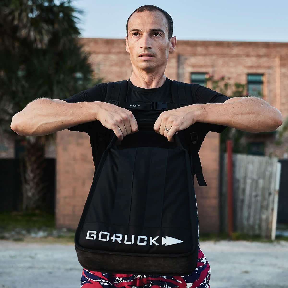 Man adjusting black GORUCK rucking gear outdoors with brick building and palm tree background