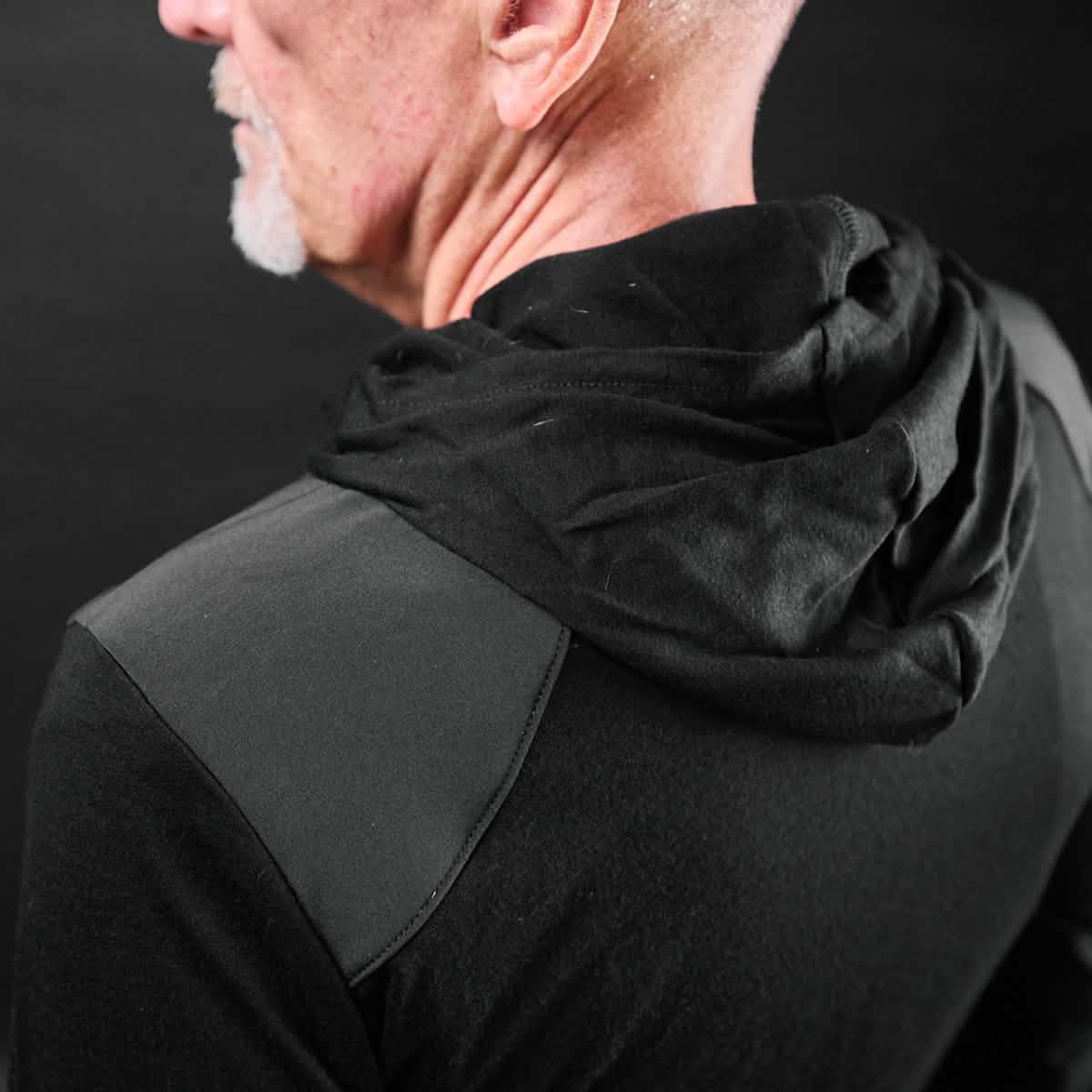 A man with a shaved head and gray beard is wearing a black Men’s Commando Pullover - Merino Wool by GORUCK, featuring a hood, viewed from the back against a dark background.