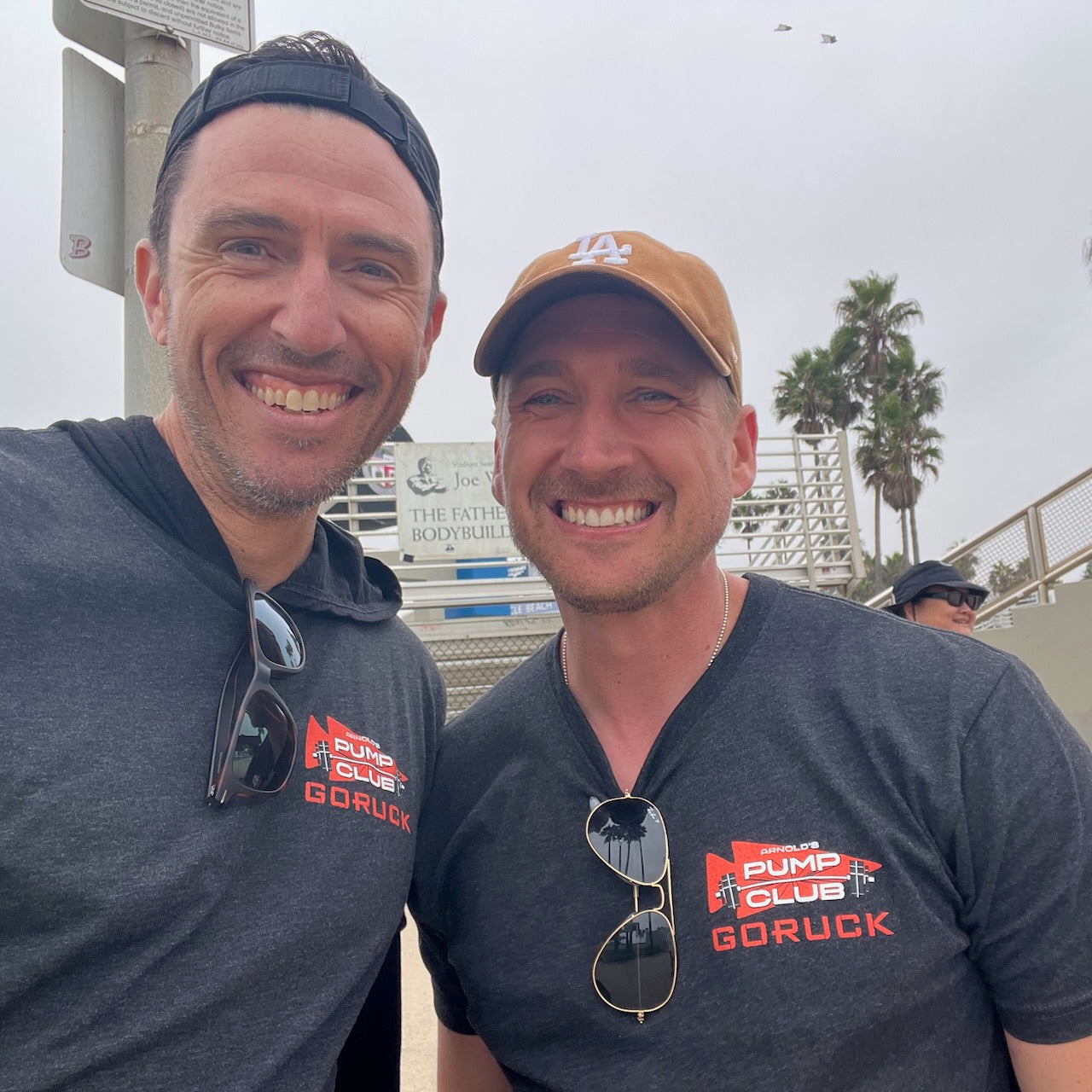 Two smiling men in matching Arnold Pump Club Tee – Tri-Blend shirts pose outdoors by a palm tree and gym equipment.