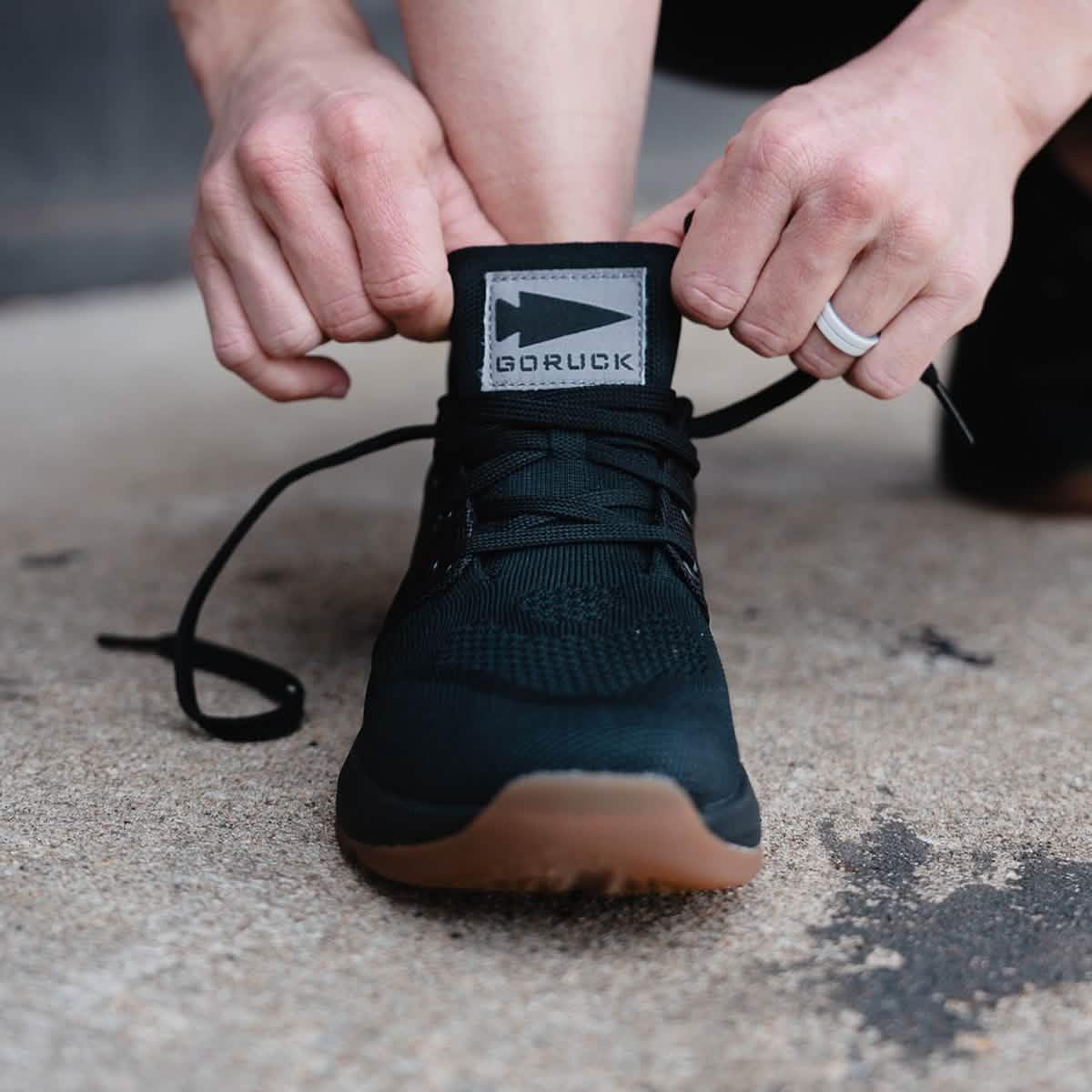 Person tying laces on a black GORUCK rucking shoe with gum sole outdoors