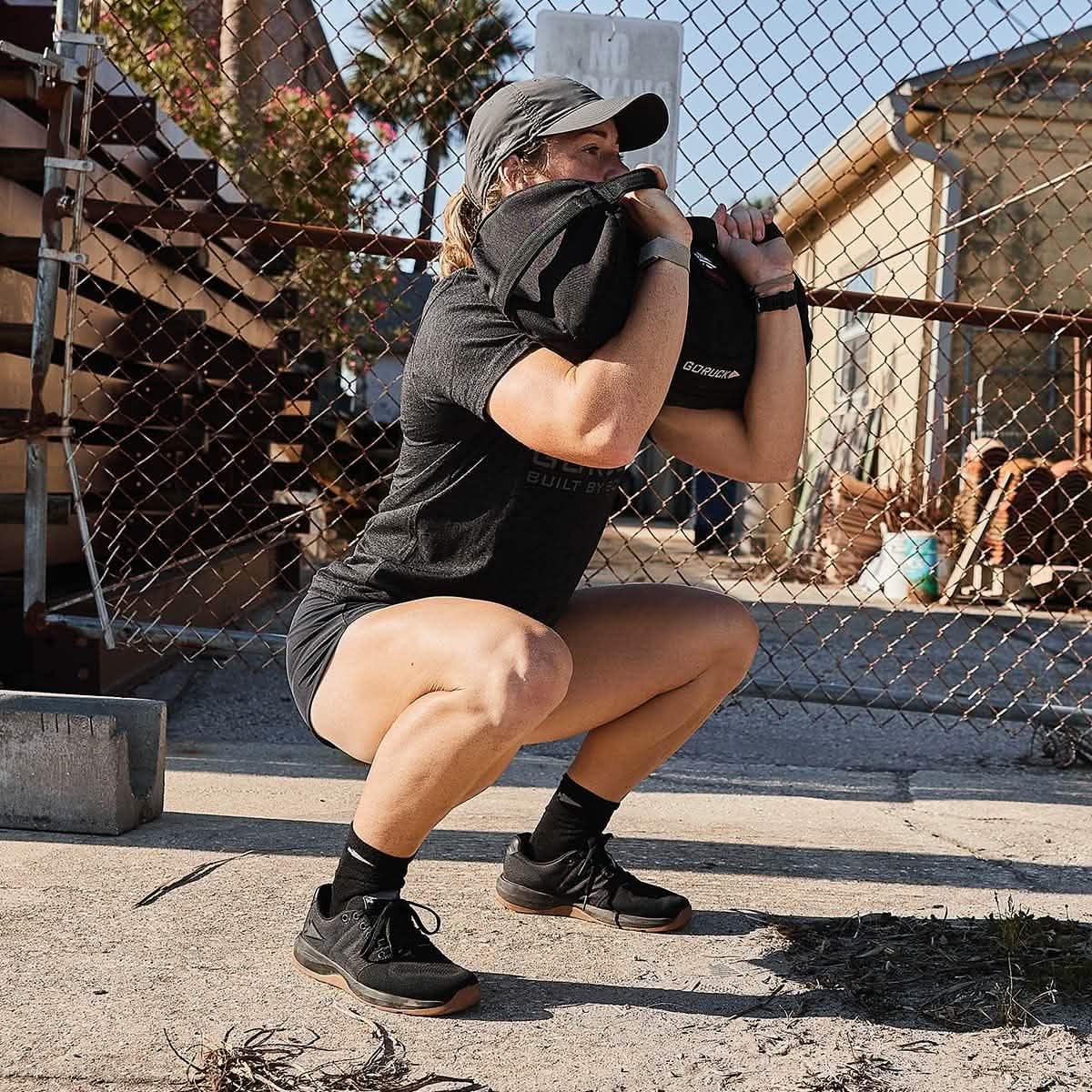 Woman performing weighted squat outdoors holding black GORUCK bag near chain-link fence