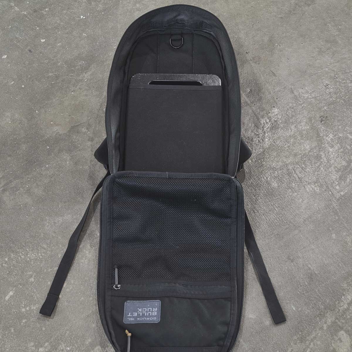 Open black backpack with a tablet or laptop inside, lying on a gray concrete floor.