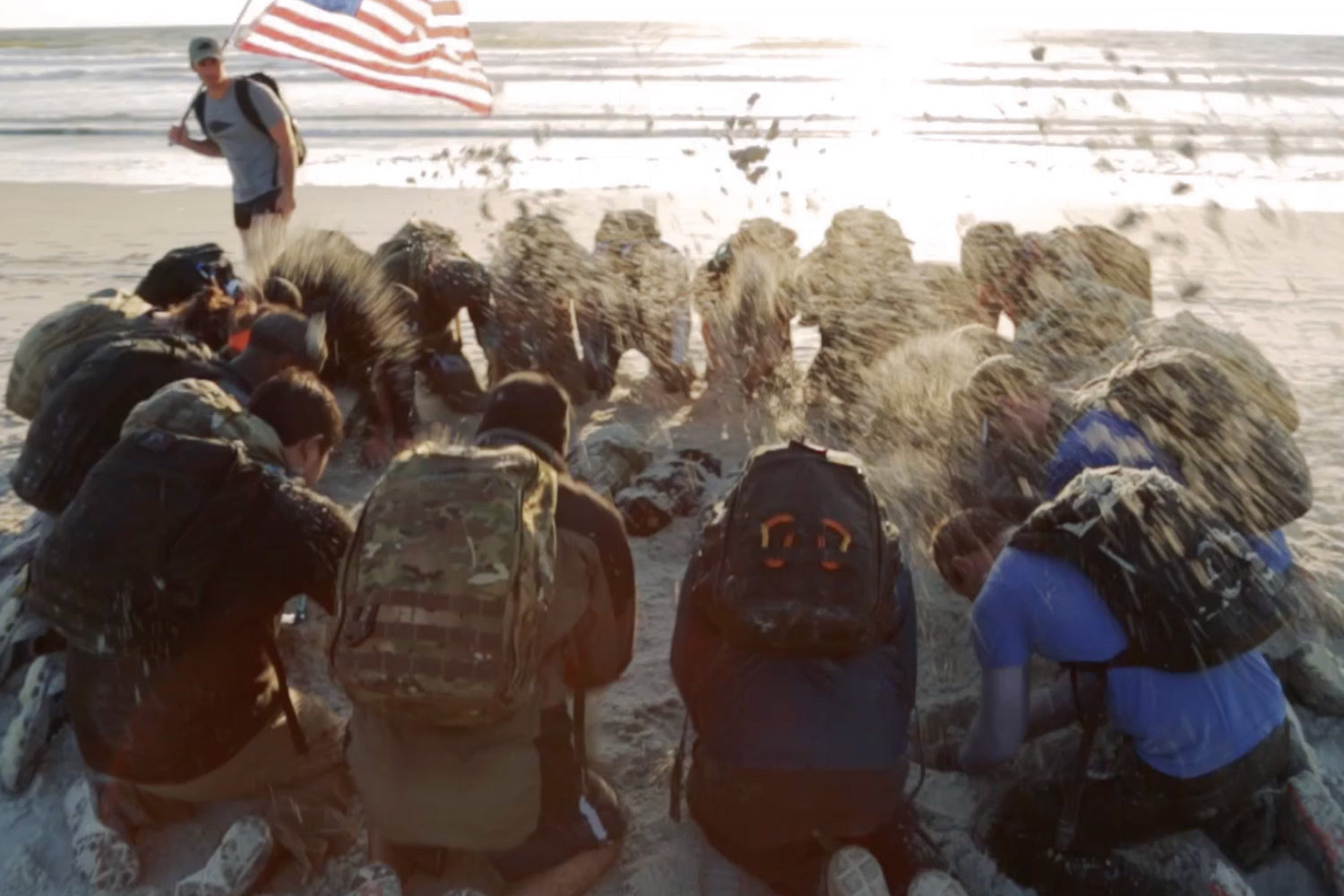 People with backpacks kneel on a sandy beach, flinging sand as a person stands behind them holding an American flag.