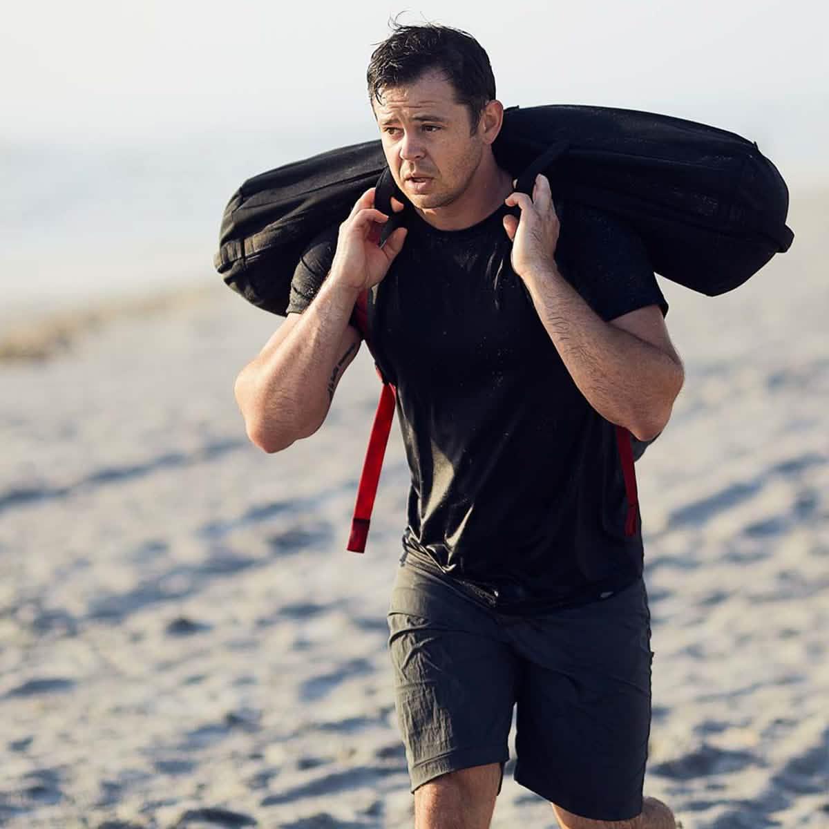 Man training on beach with GORUCK sandbag, wearing black athletic gear