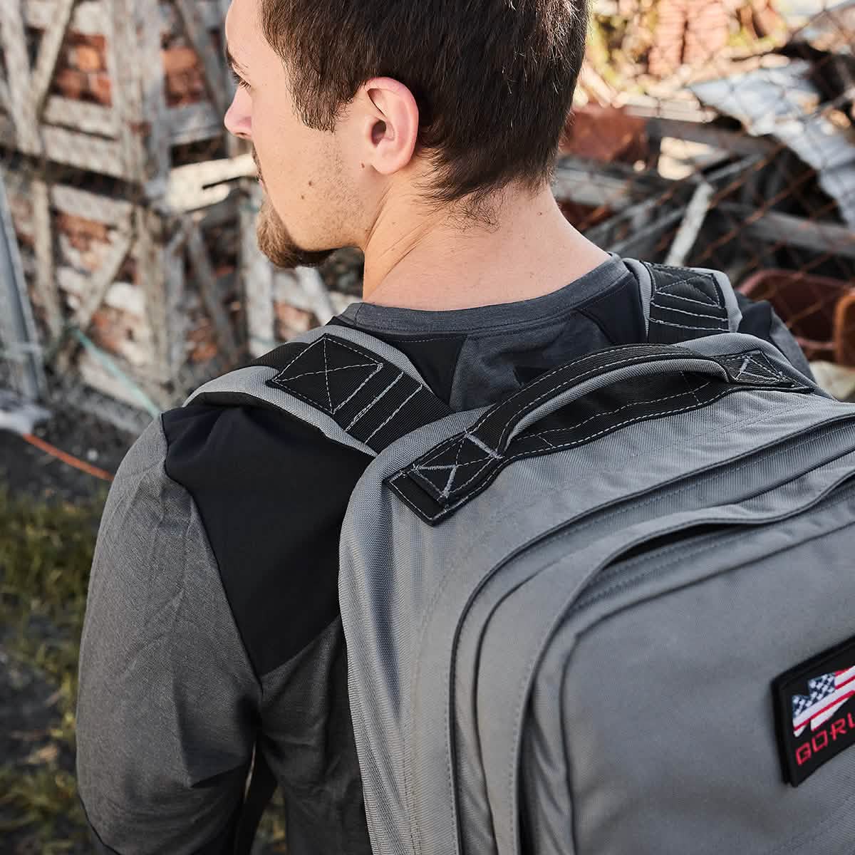 A man with brown hair and a goatee, wearing a GORUCK Men’s Commando Long Sleeve made of Merino Wool, strolls with a gray backpack. He is outdoors near a fence, surrounded by wood and brick debris in the background.