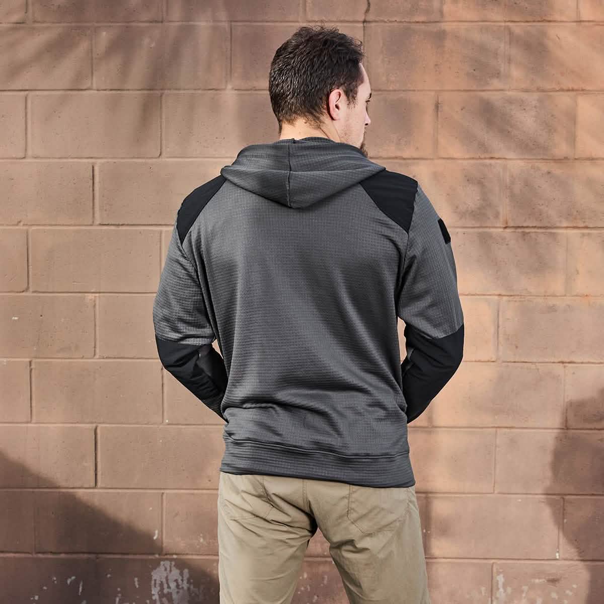 A man stands with his back to the camera, wearing the Men’s Full Zip - Polartec Grid Fleece from GORUCK in a gray color with black accents and khaki pants. He is positioned in front of a textured, light brown brick wall that features faint tree branch shadows.