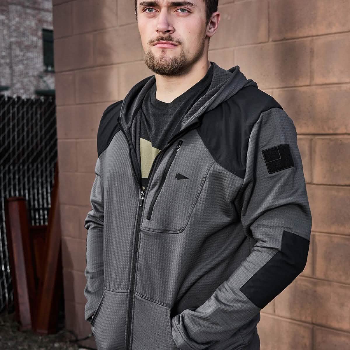 A man with a beard stands outdoors against a brick wall, wearing the GORUCK Men’s Full Zip - Polartec Grid Fleece in gray and black. The fleece-lined jacket features a hood, complementing his rugged look against the backdrop of a chain-link fence and industrial elements.