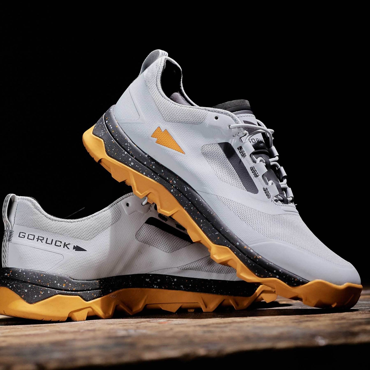 A pair of Mackall trail running shoes in Glacier and Ignition Orange, featuring a triple compound outsole ideal for rucking, is displayed on a wooden surface against a black background. The brand name GORUCK is visible on the heel.