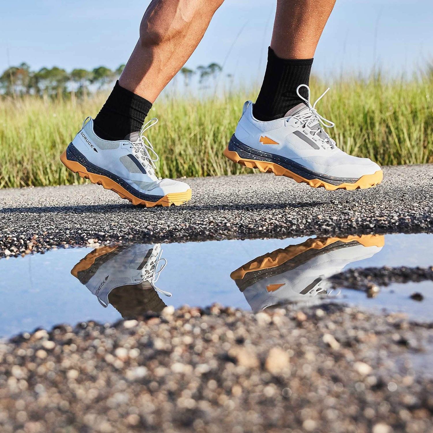 A person running on a paved path beside a puddle is wearing GORUCK's Mackall shoes in Glacier and Ignition Orange, featuring a triple compound outsole, along with black socks. The green grass and trees in the background are slightly blurred, highlighting the motion.