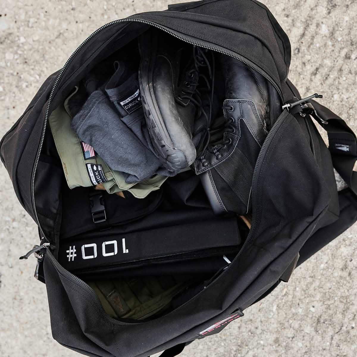 Top view of open black GORUCK duffel bag packed with tactical boots, folded GORUCK pants, and rucking gear on concrete floor
