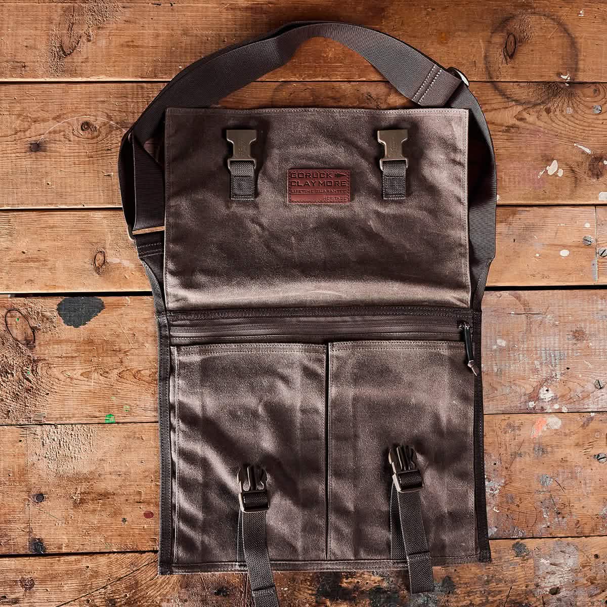 The GORUCK Heritage Claymore Sling Bag, crafted from rugged brown waxed canvas and accented with leather straps and a shoulder strap, is showcased open on a rustic wooden surface. Its design incorporates metal buckles that reflect the bag's durable, utilitarian heritage.