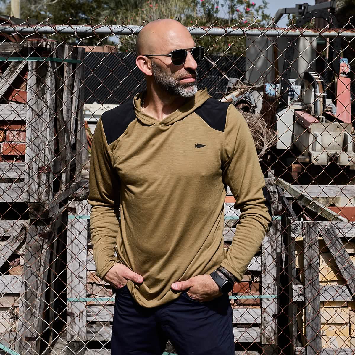 A bald man with a beard is wearing sunglasses and the GORUCK Men’s Commando Pullover - Merino Wool in tan, which features reinforced paneling on the shoulders. He stands in front of a chain-link fence with stacks of wooden pallets in the background, under a clear sky.