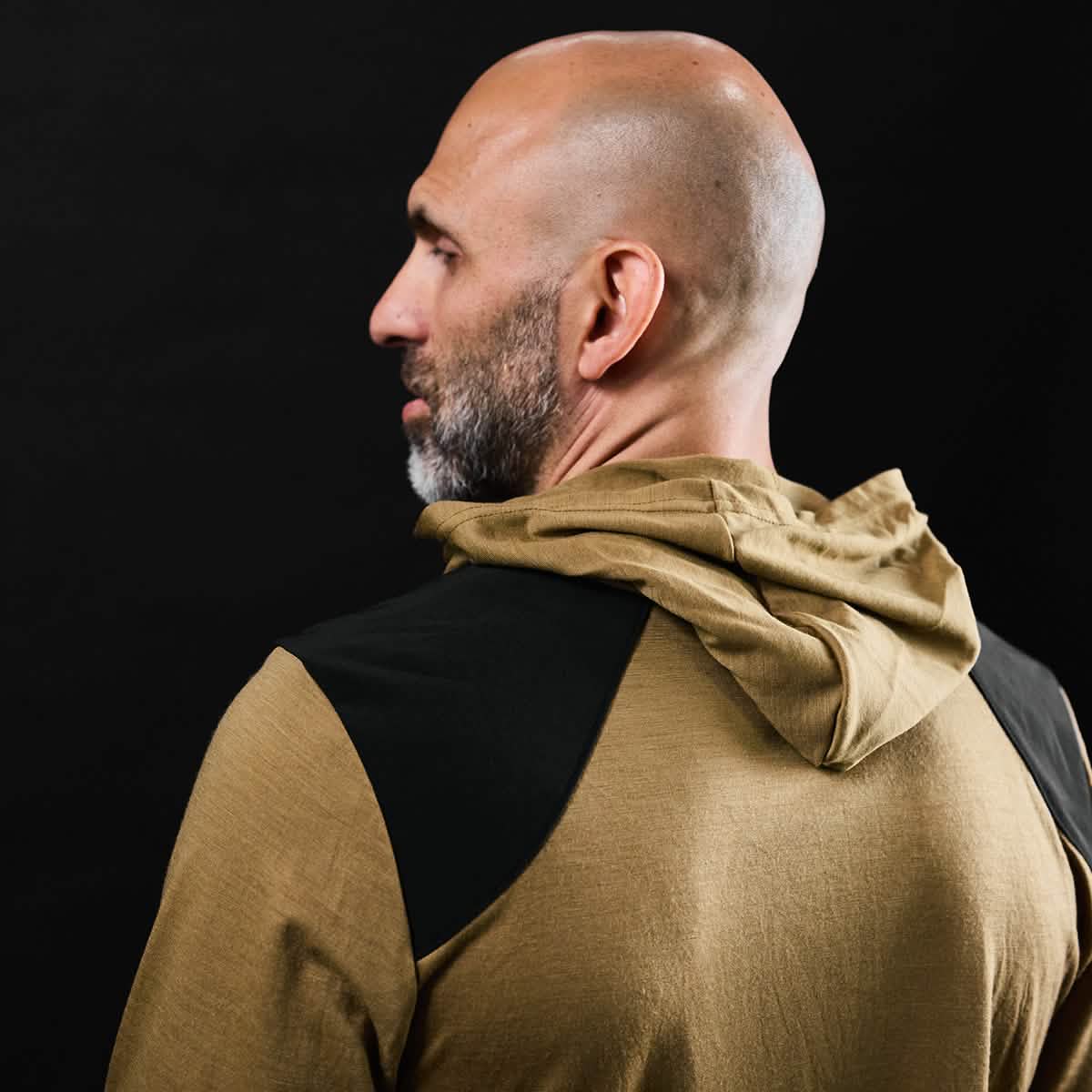 A bald man with a beard is turned to the side, wearing a GORUCK Men’s Commando Pullover made from merino wool, featuring black shoulder accents reminiscent of a British Commando Sweater, set against a dark background.