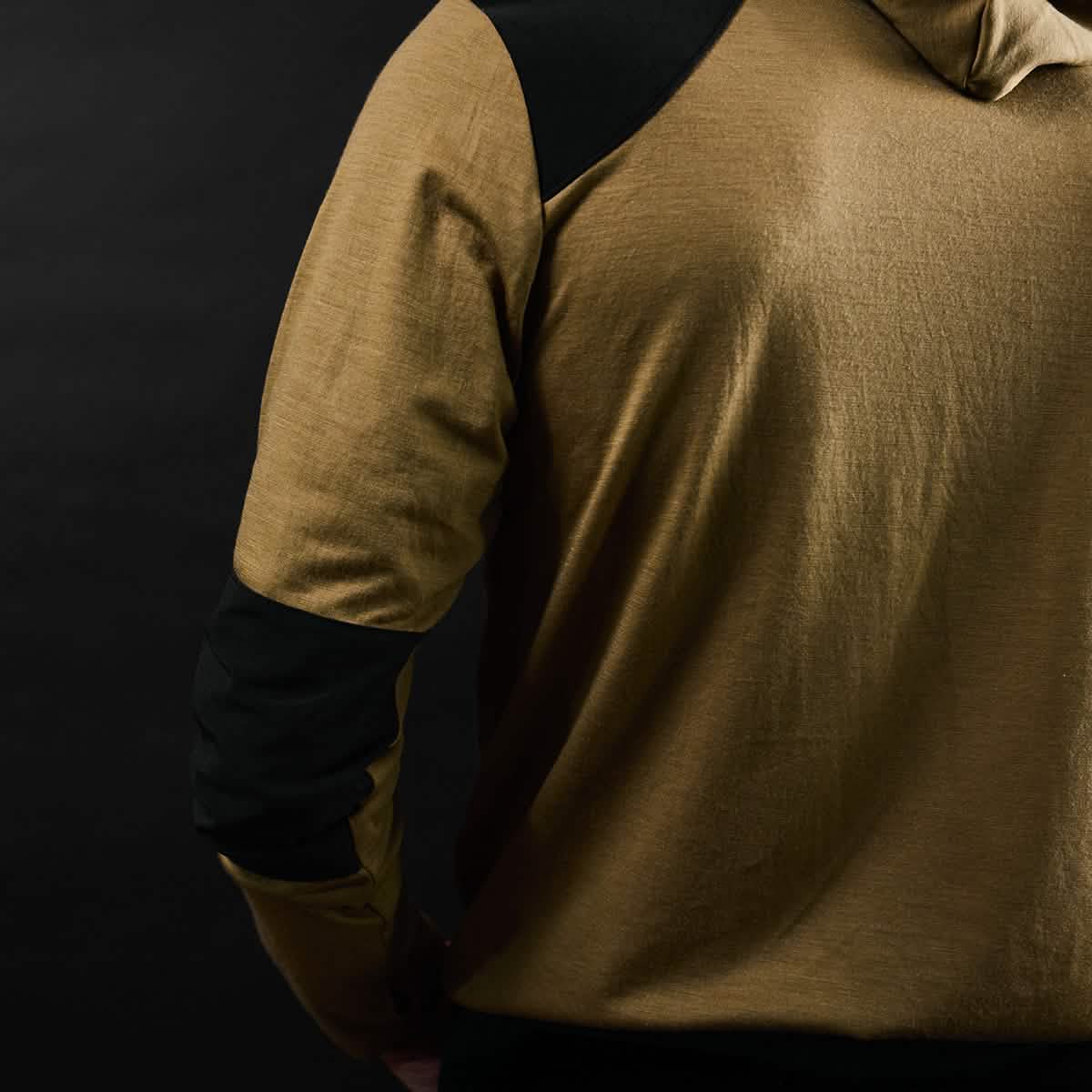 A person wearing the Men’s Commando Pullover - Merino Wool by GORUCK, featuring tan fabric with black shoulder and elbow patches, stands against a dark background.
