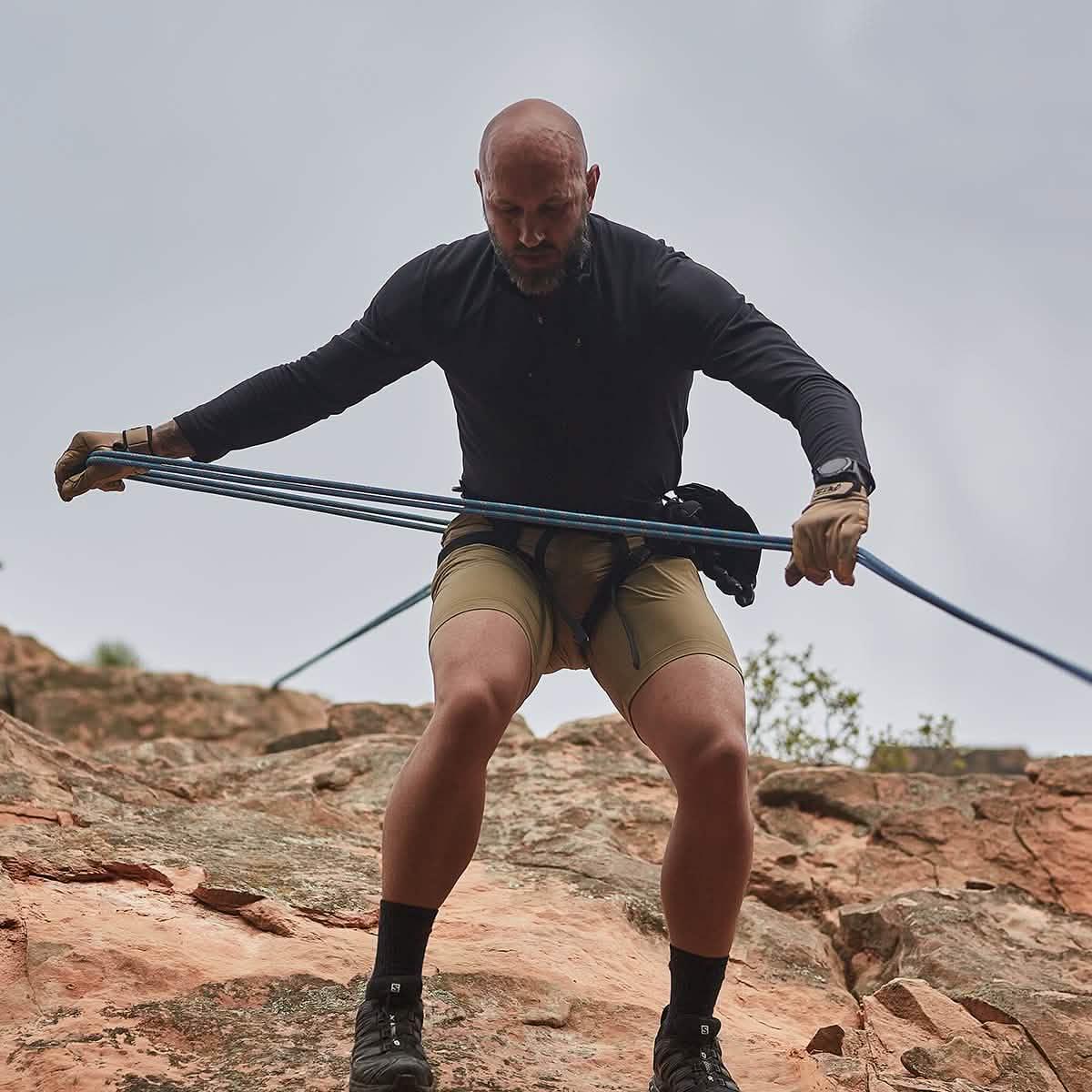 Man in GORUCK gear rappelling down a rocky cliff using blue rope outdoors