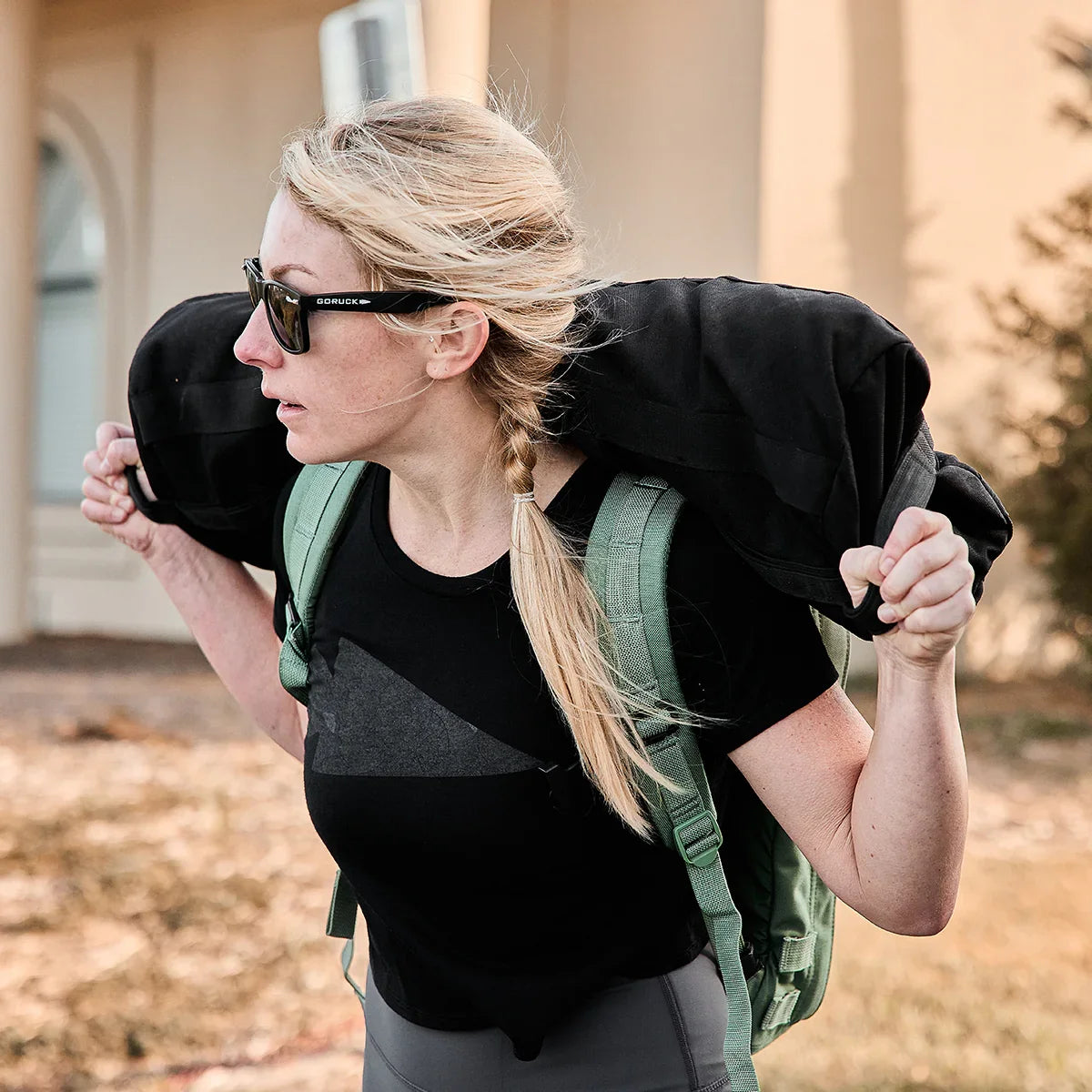 Woman wearing GORUCK sunglasses and green backpack carrying black rucking sandbag outdoors