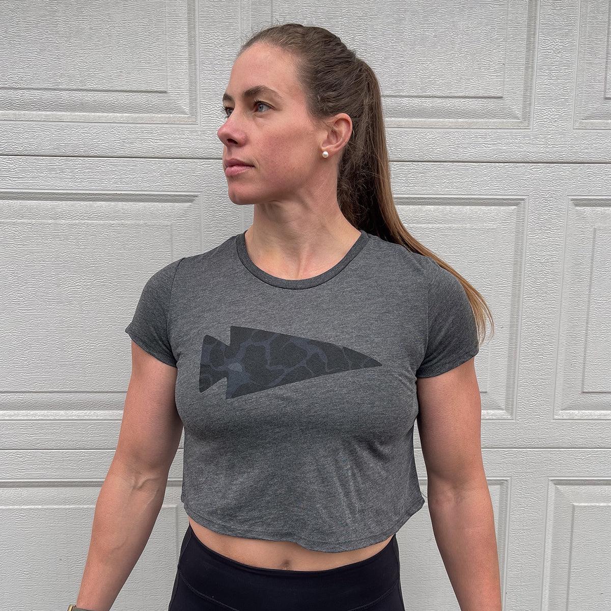 fit woman in grey cropped GORUCK t-shirt with dark arrowhead logo standing against textured garage door