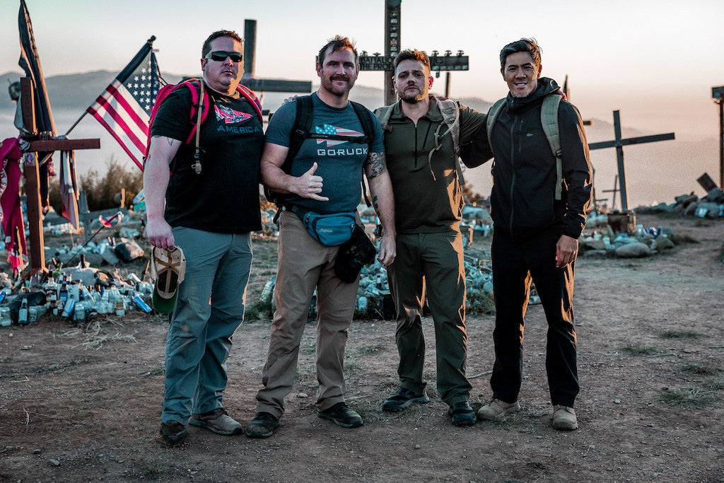 Four men wearing GORUCK rucking gear posing at a memorial site with crosses and an American flag at sunset