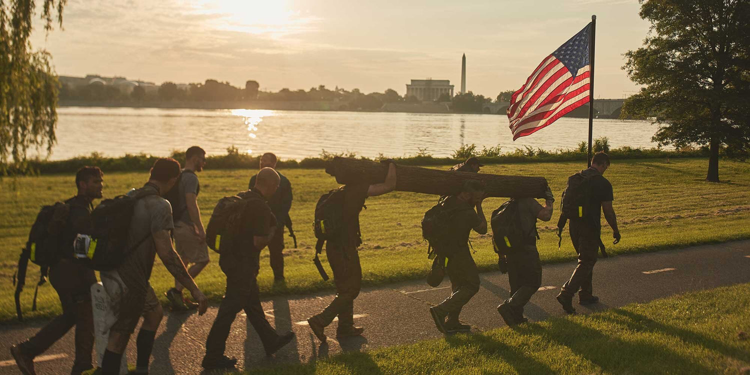 A group of people carry a log and an American flag along a riverside path at sunrise.